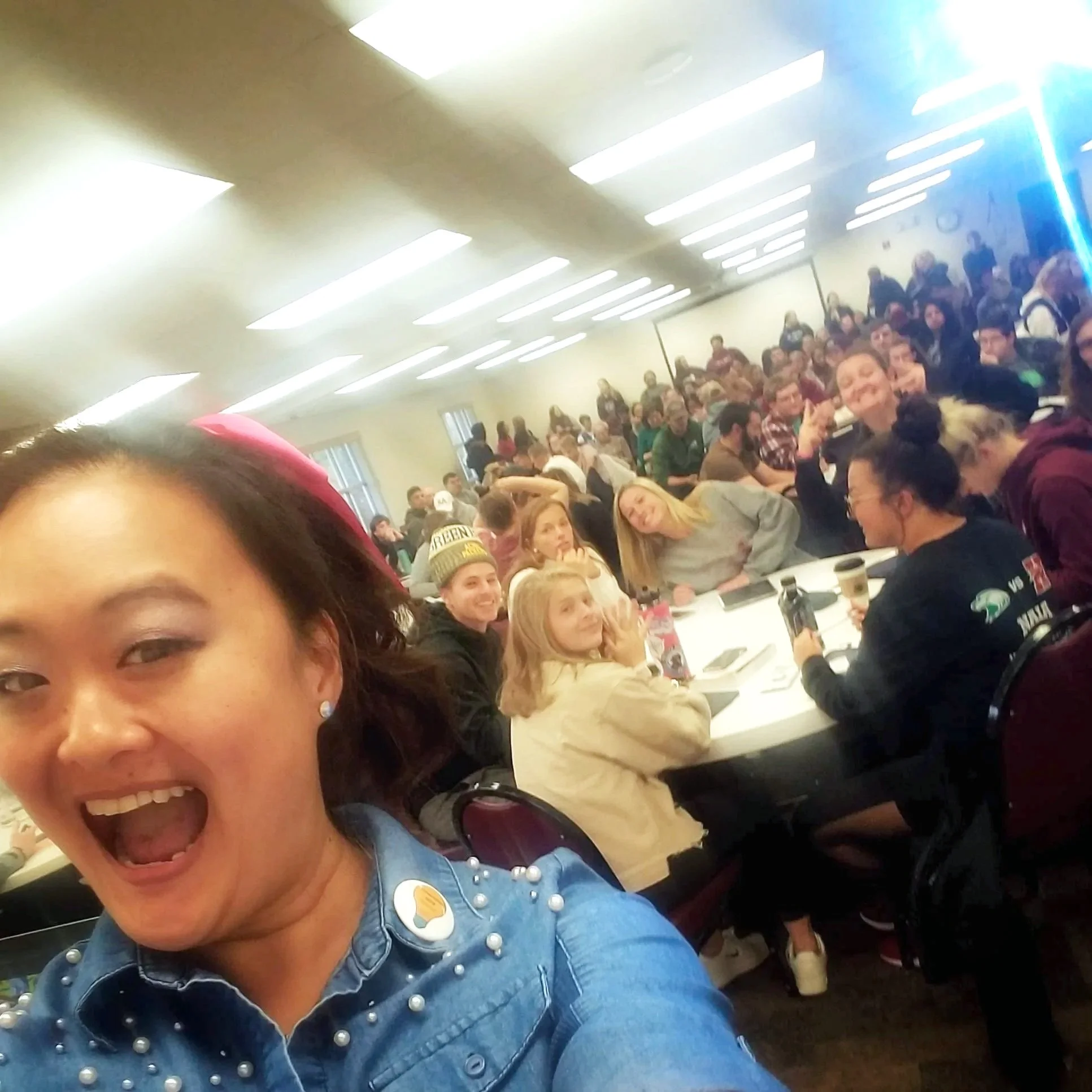 Mary Bowers, wearing a blue denim shirt with pearl embellishments, is smiling and laughing in the foreground. She is joined by many students from Hastings College, who are seated at tables in the background.