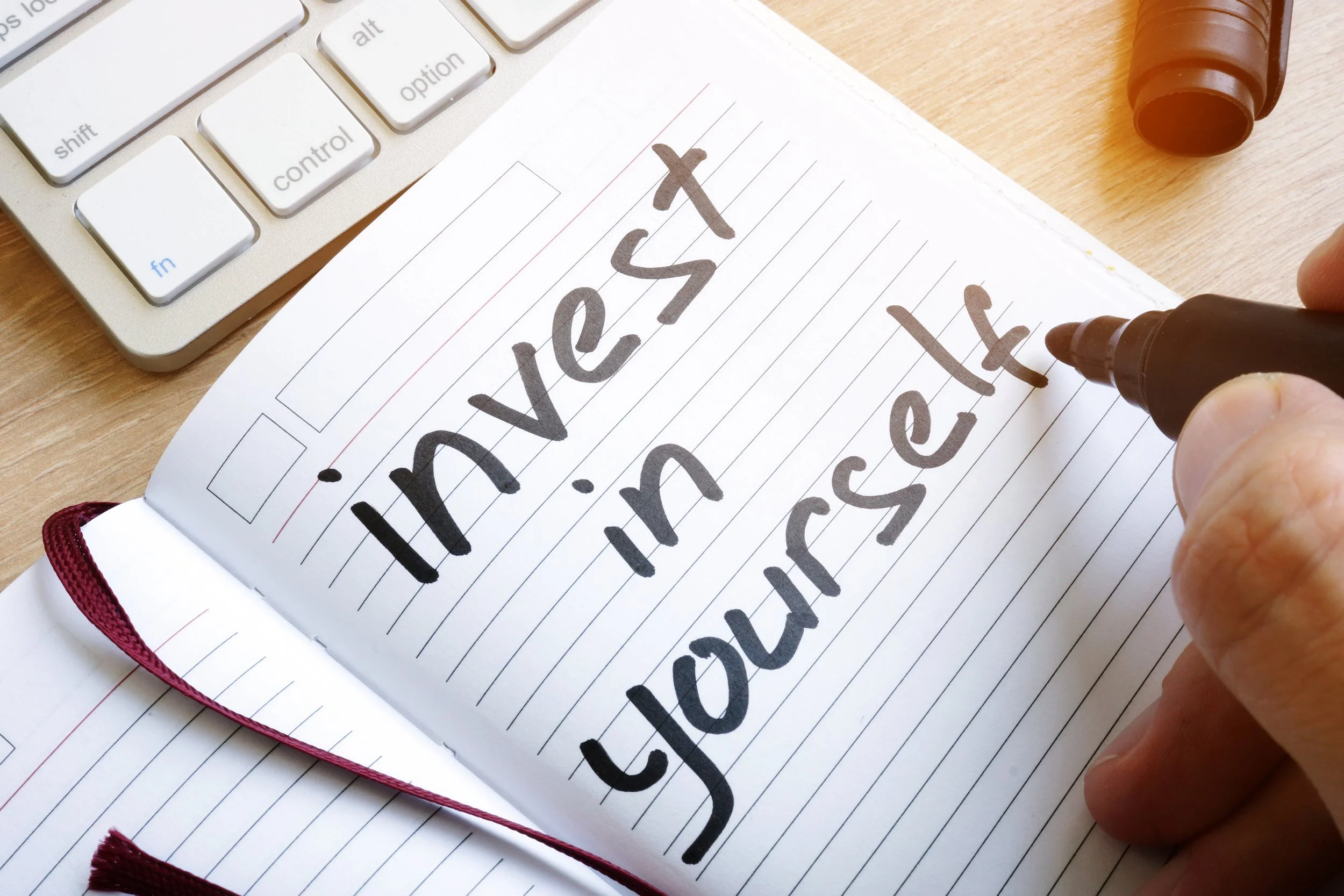 Close-up of a handwritten journal page with the phrase "I lovet you" written in bold black marker, with a person's hand holding a black marker, near a computer keyboard and a brown glue stick on a wooden surface.