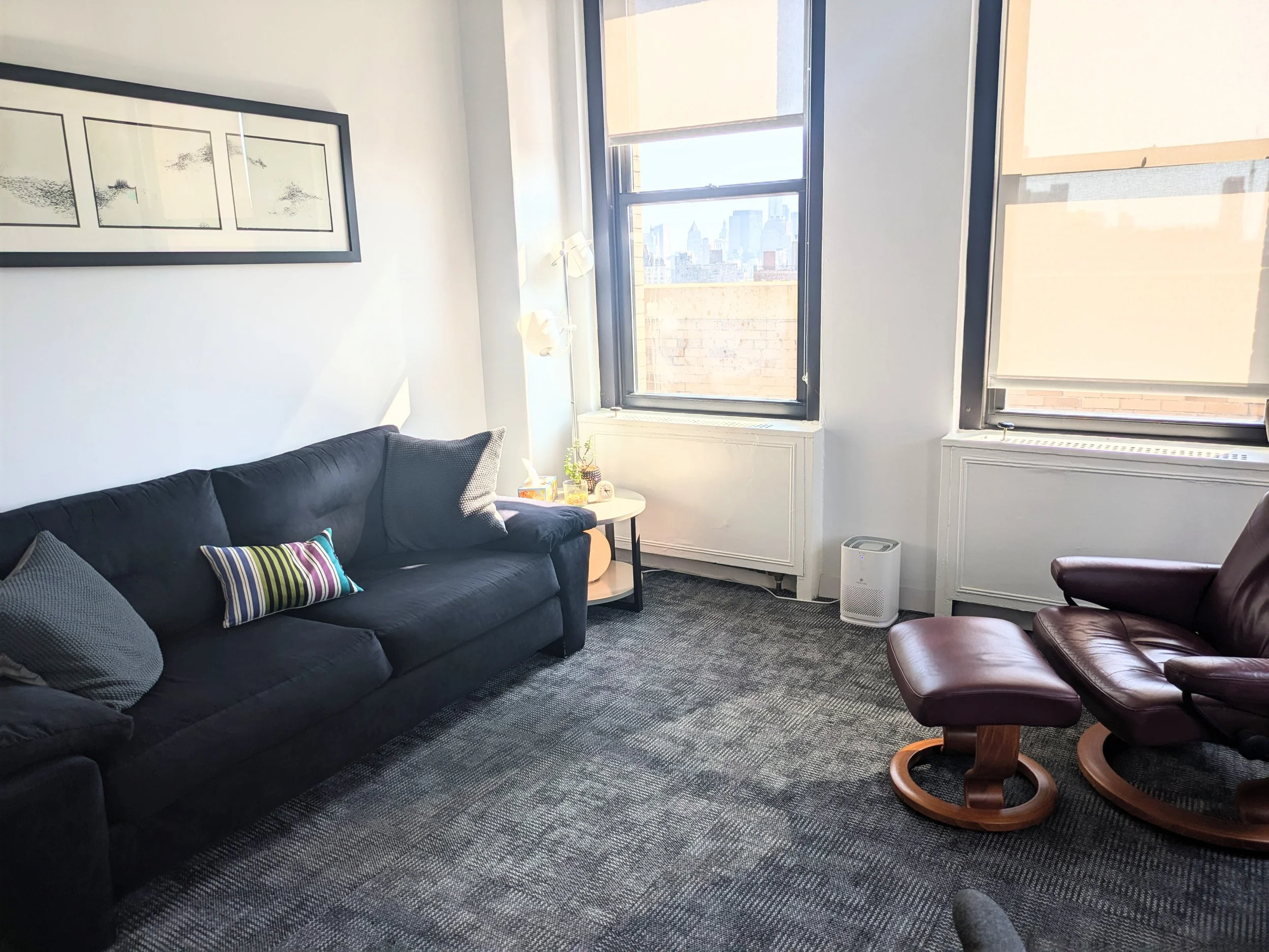 A living room with a black sofa decorated with multiple colorful pillows, chair with footrest, and windows with city skyline. An in-person therapy room in Manhattan for therapy sessions with adults, couples, and teenagers in New York.