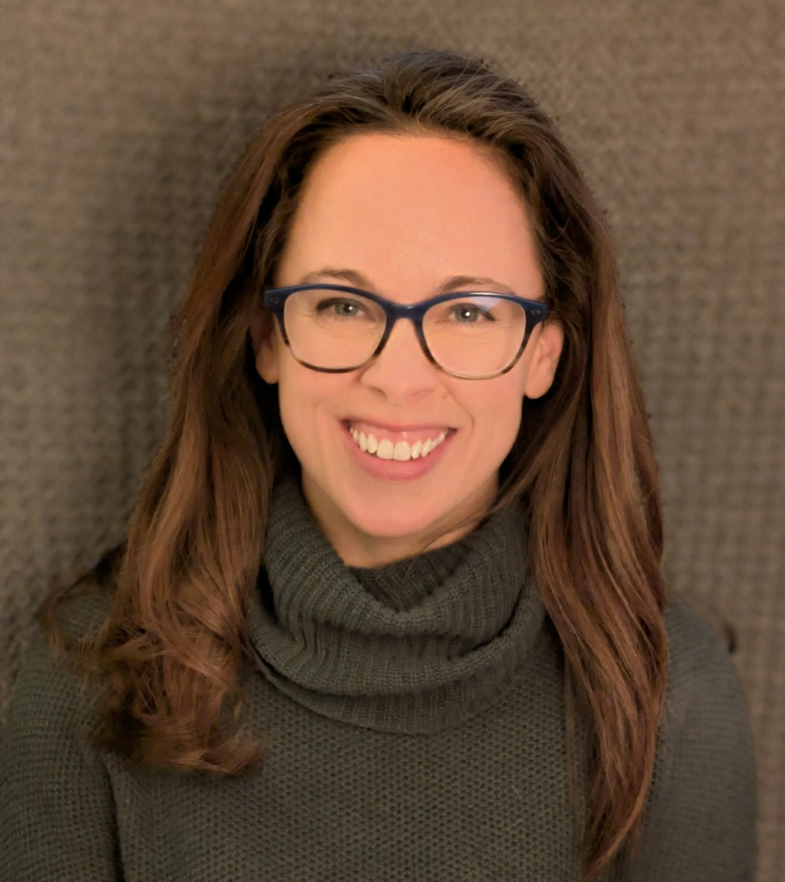 Close-up of a white female therapist with long reddish-brown hair, wearing glasses with a dark frame, smiling, and dressed in a gray, textured sweater, against a brownish background.