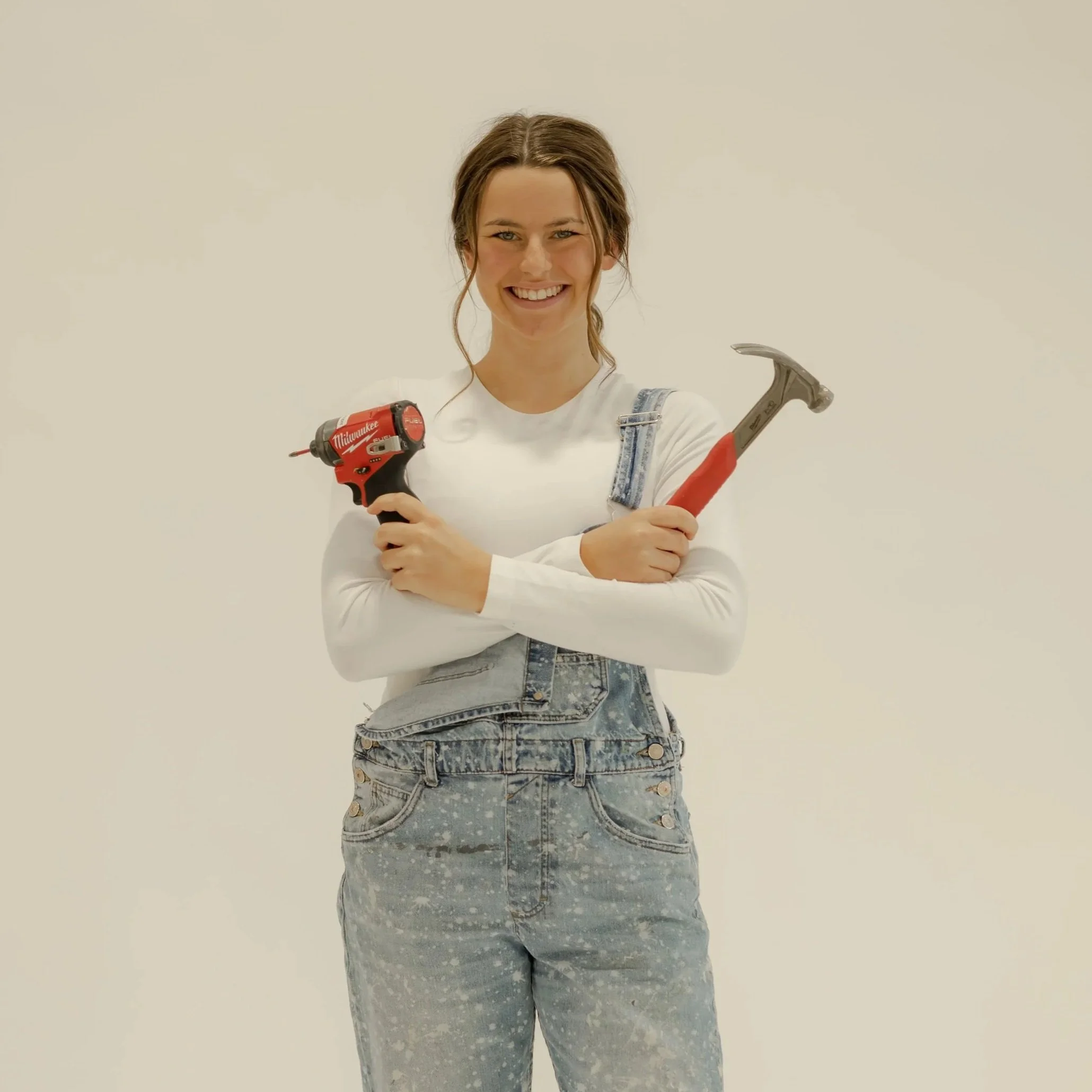 Smiling woman holding a cordless power drill and a hammer, wearing a white shirt and distressed denim overalls, standing against a plain background.