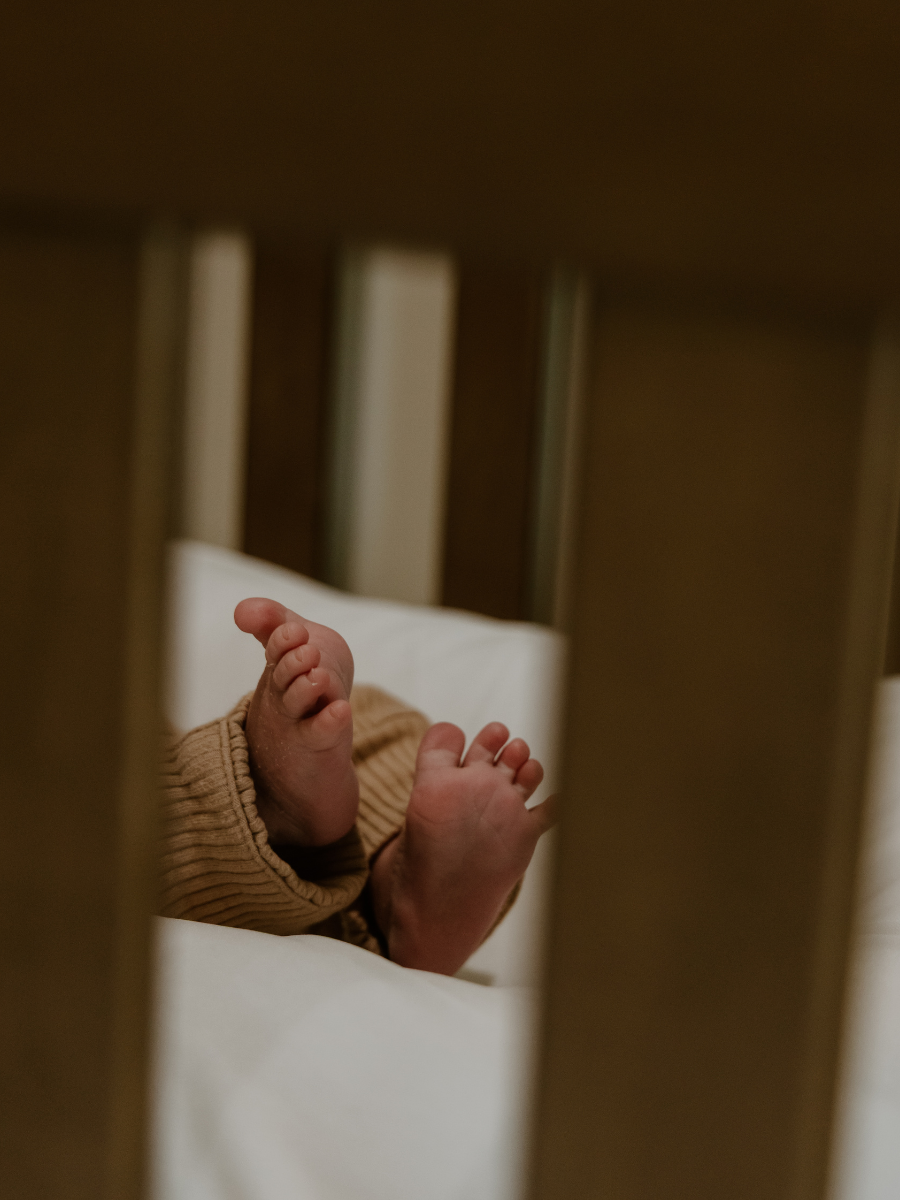 View through crib slats showing a baby's feet with toes curled, wearing a tan-colored ribbed sock or clothing.
