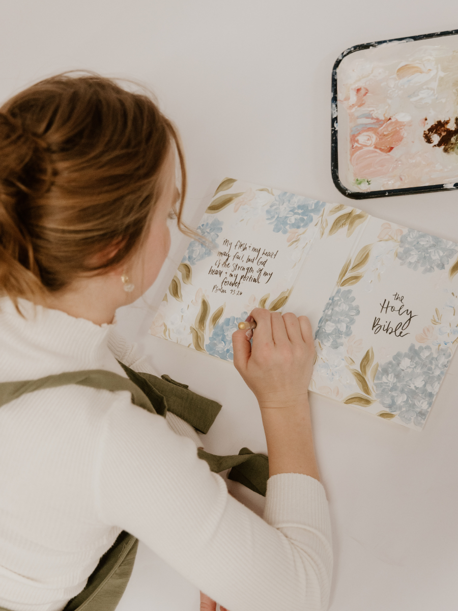A woman with brown hair in a white sweater writes in a floral decorated Bible, sitting at a white table with a paint palette nearby.
