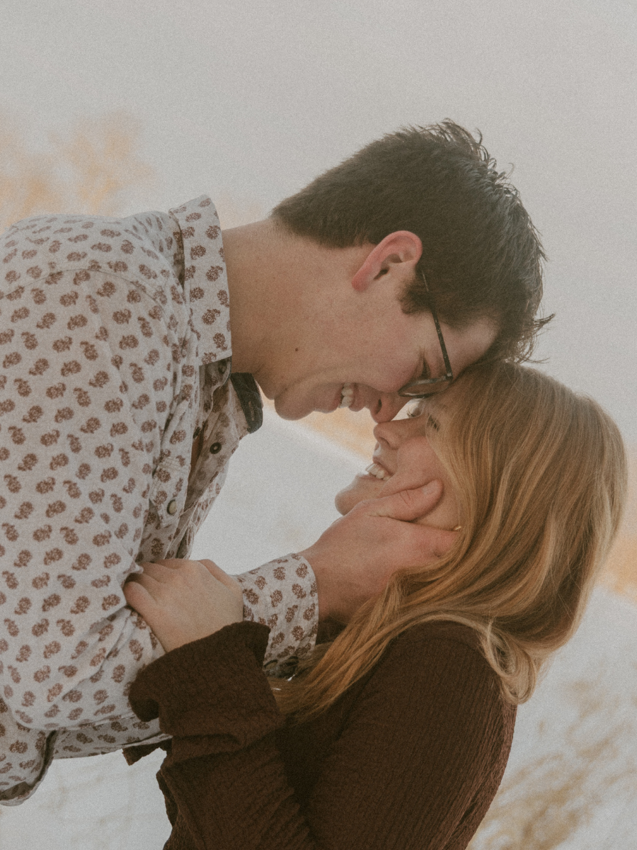 A man and woman are sharing a joyful moment, smiling and touching each other's faces, indoors with a neutral background.