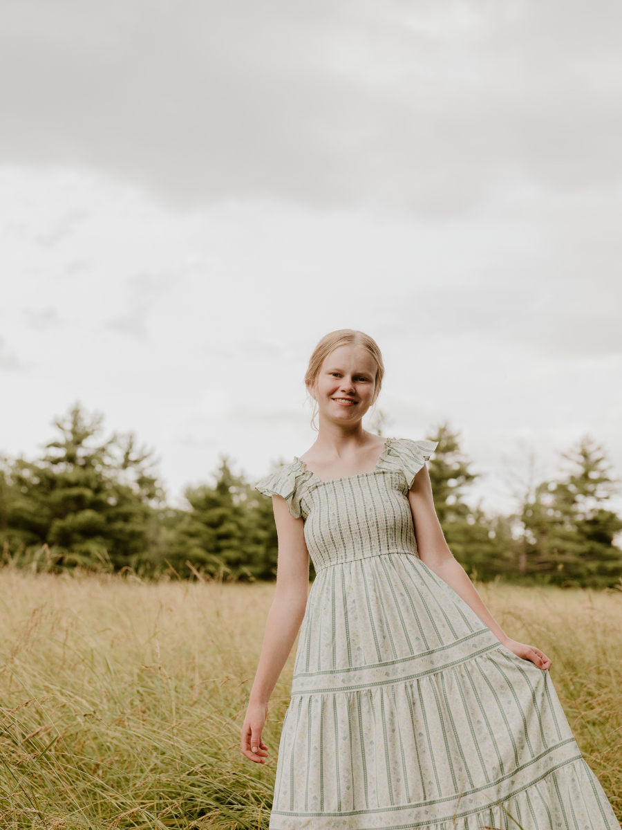 A young girl with blonde hair wearing a light-colored, striped, flowy dress standing in a grassy field with trees in the background under a cloudy sky.