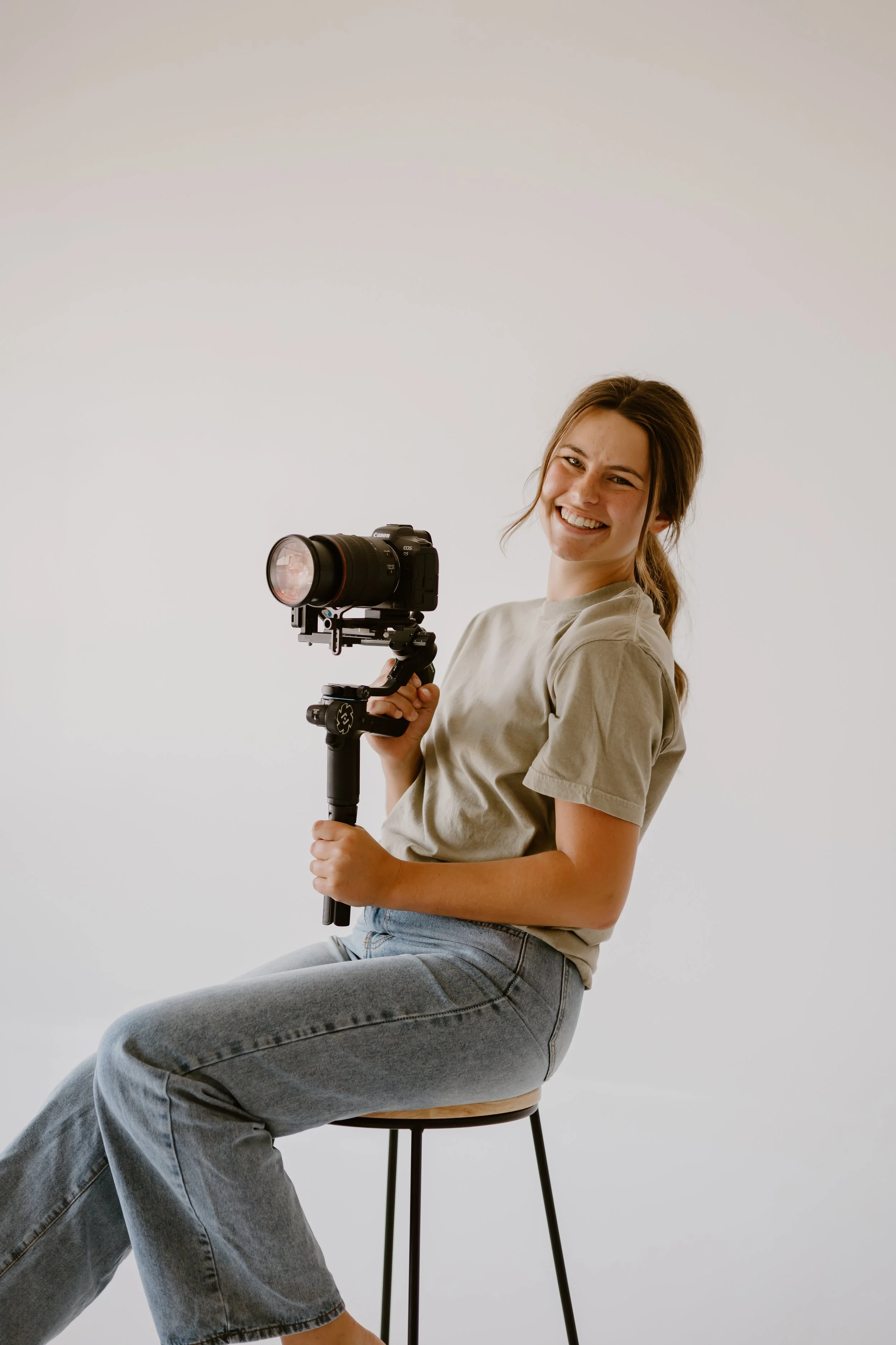 A young woman smiling and holding a camera with a tripod rig, sitting on a stool against a plain white background.