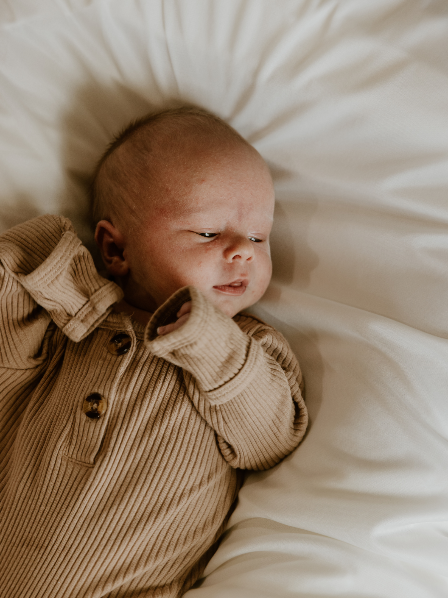 A baby lying on a white pillow, wearing a brown ribbed button-up shirt, with a serious expression.