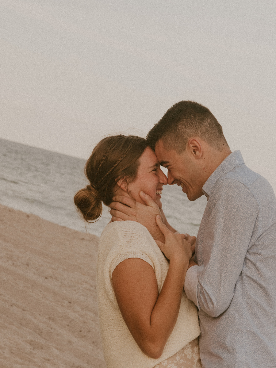 A couple standing close together on a beach, smiling and touching foreheads with their noses almost touching, with ocean waves in the background.