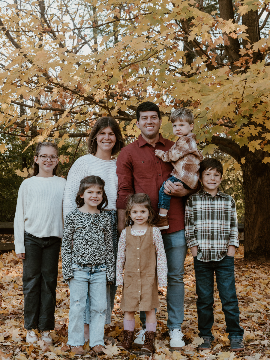 Family of seven posing outdoors in autumn, standing on fallen leaves under a large tree with yellow and orange leaves, smiling at the camera.