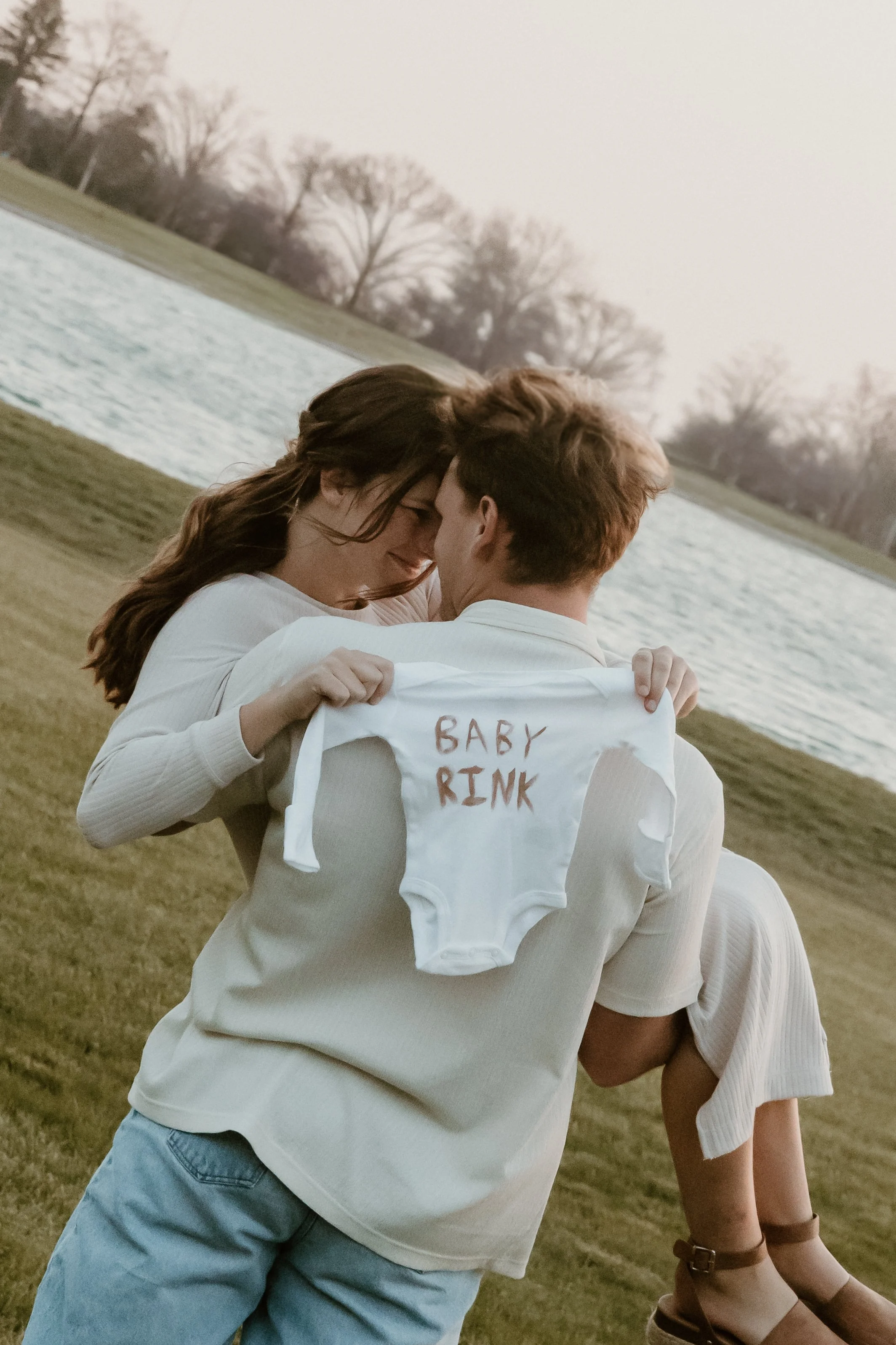 A couple hugging outdoors, with a baby onesie that reads 'Baby Pink' hanging from one person's back, near a pond and leafless trees in the background.