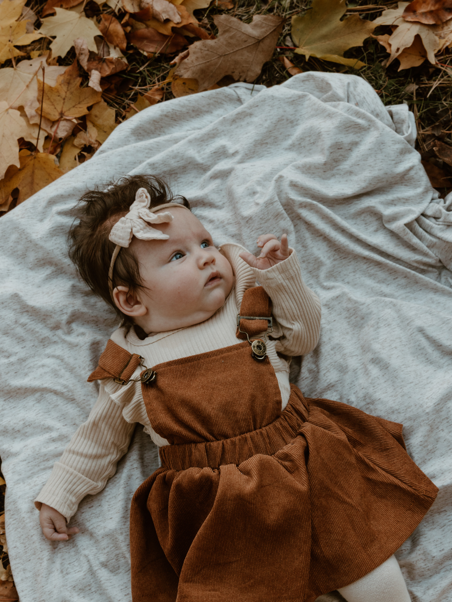 A baby girl with a beige bow headband lying on a blanket outdoors among fallen autumn leaves, wearing a beige long-sleeve shirt and brown corduroy overalls.