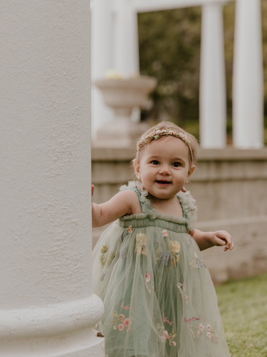 Young girl peeking out from behind a white pillar outdoors in a dress with embroidered flowers, smiling at the camera.