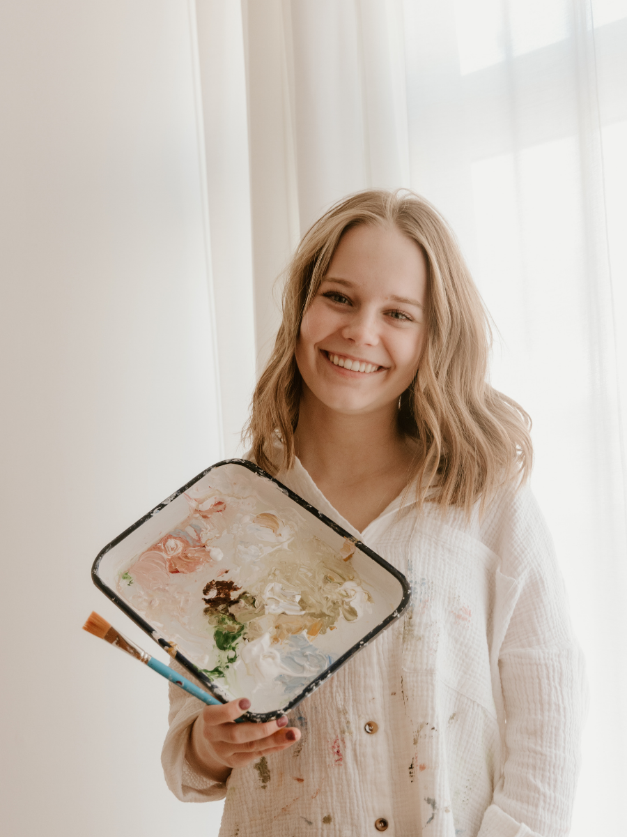 A young woman with blonde hair holding a paint palette with various colors and a paintbrush, smiling indoors near a window with sheer curtains.