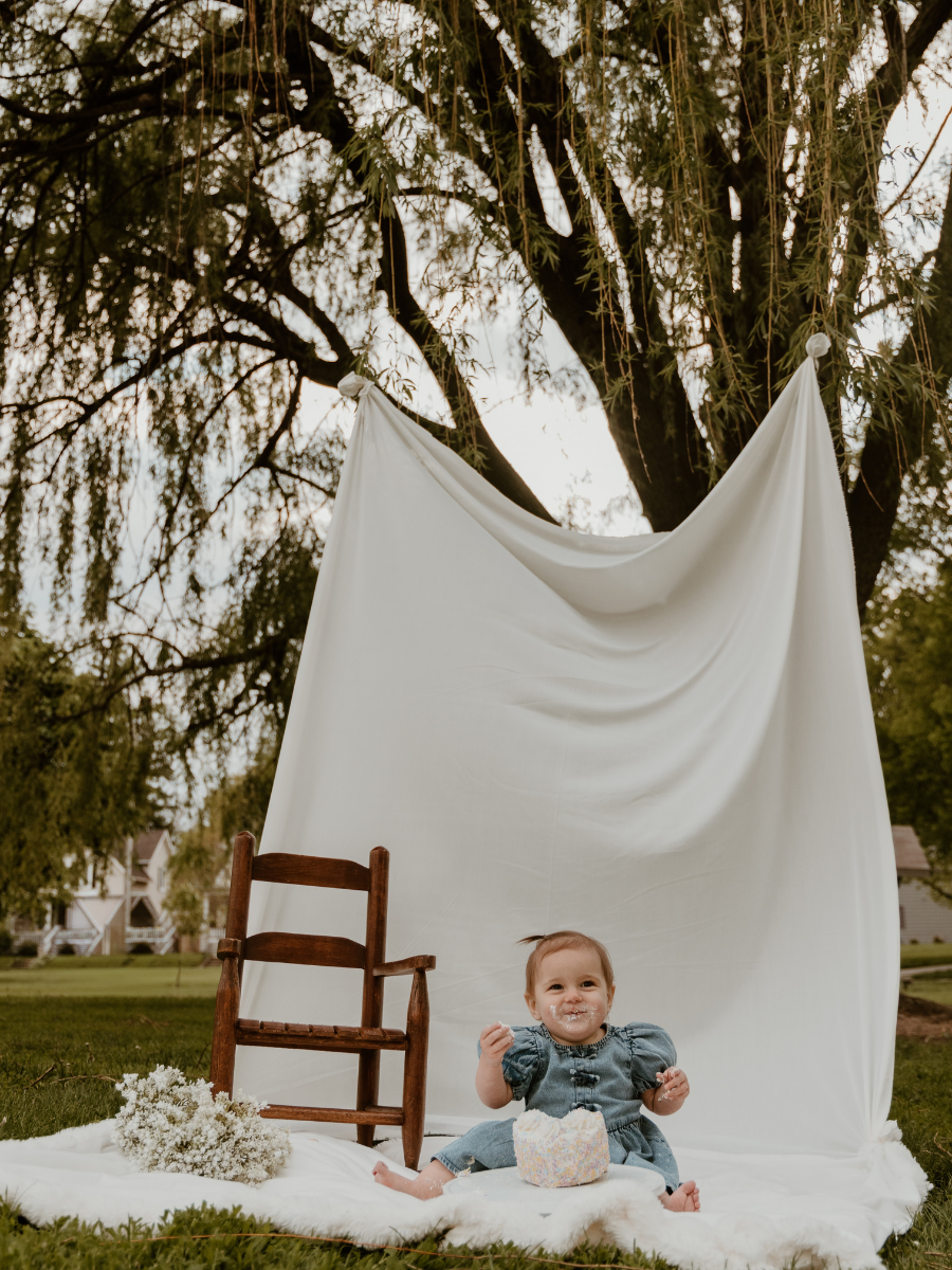 A baby girl sitting on a white blanket outdoors with a white fabric backdrop hanging from a tree. The baby has cake on her face and around her mouth, holding a piece of cake, with a small cake nearby. There's a small wooden chair and a bunch of white