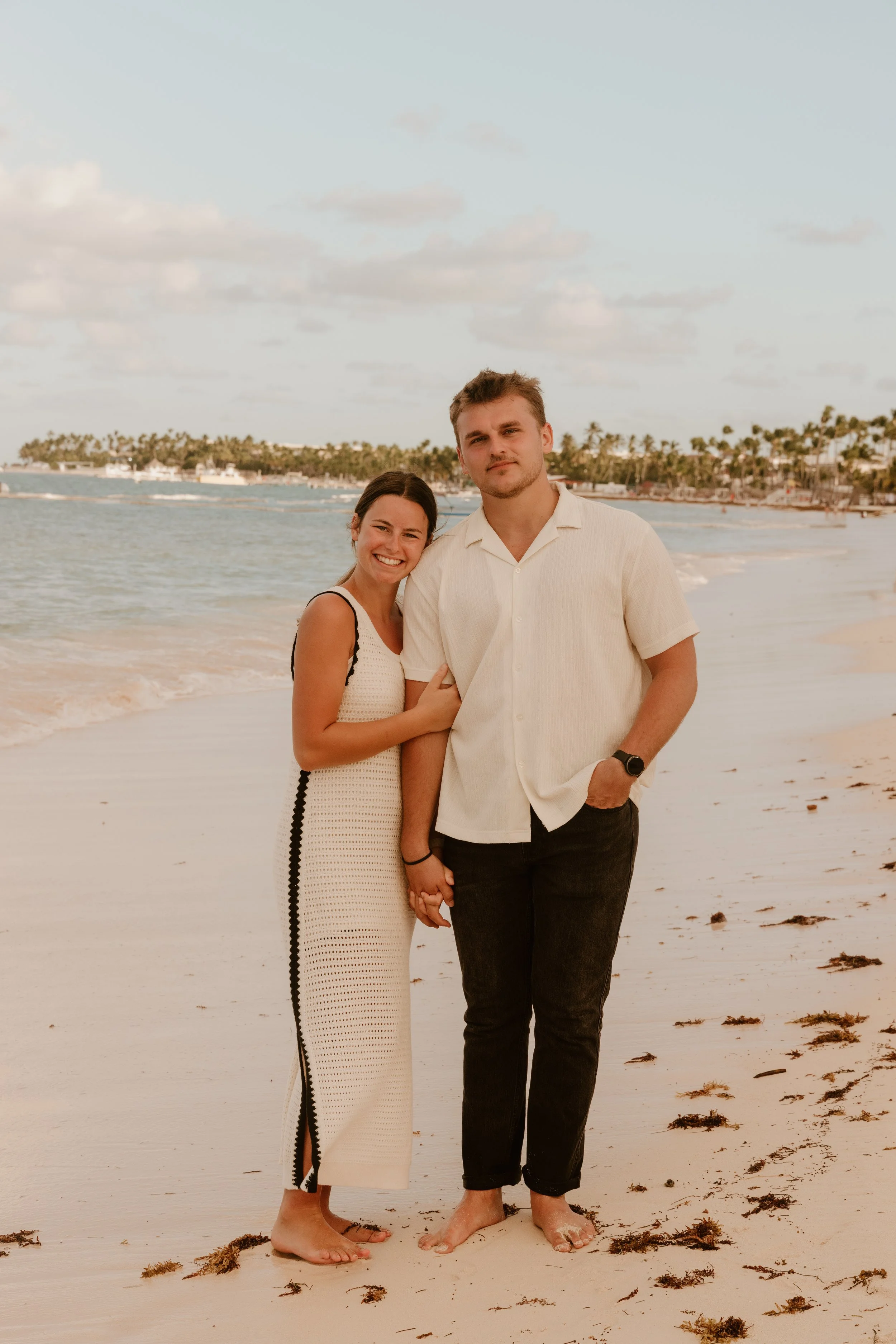 A smiling woman and a man stand on a beach, holding hands, with the woman leaning her head on the man's shoulder. The woman is wearing a sleeveless white knit dress with black trim, and the man is wearing a light-colored short-sleeve shirt and black pants. The background features the ocean, a partly cloudy sky, and a distant shoreline with palm trees.