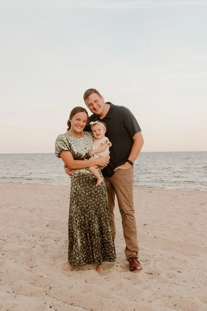 A happy family of three standing on a sandy beach near the ocean at sunset, smiling and embracing each other.