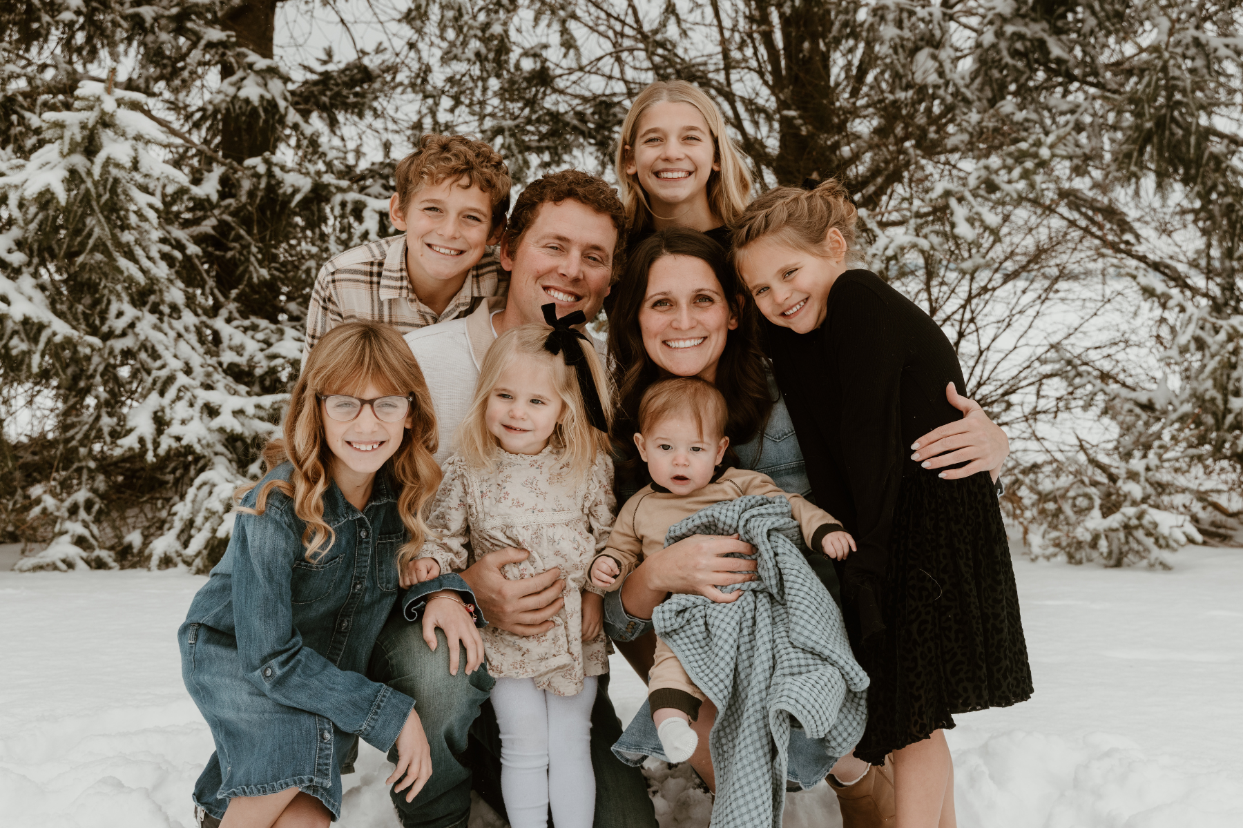 A large family group of ten people, including children and adults, smiling and huddled together outdoors in the snow with snow-covered trees in the background.