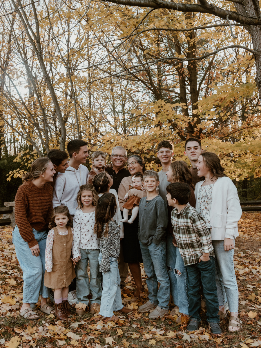 Family and friends gathering outdoors among autumn trees with fall leaves on the ground, smiling and posing for a group photo.