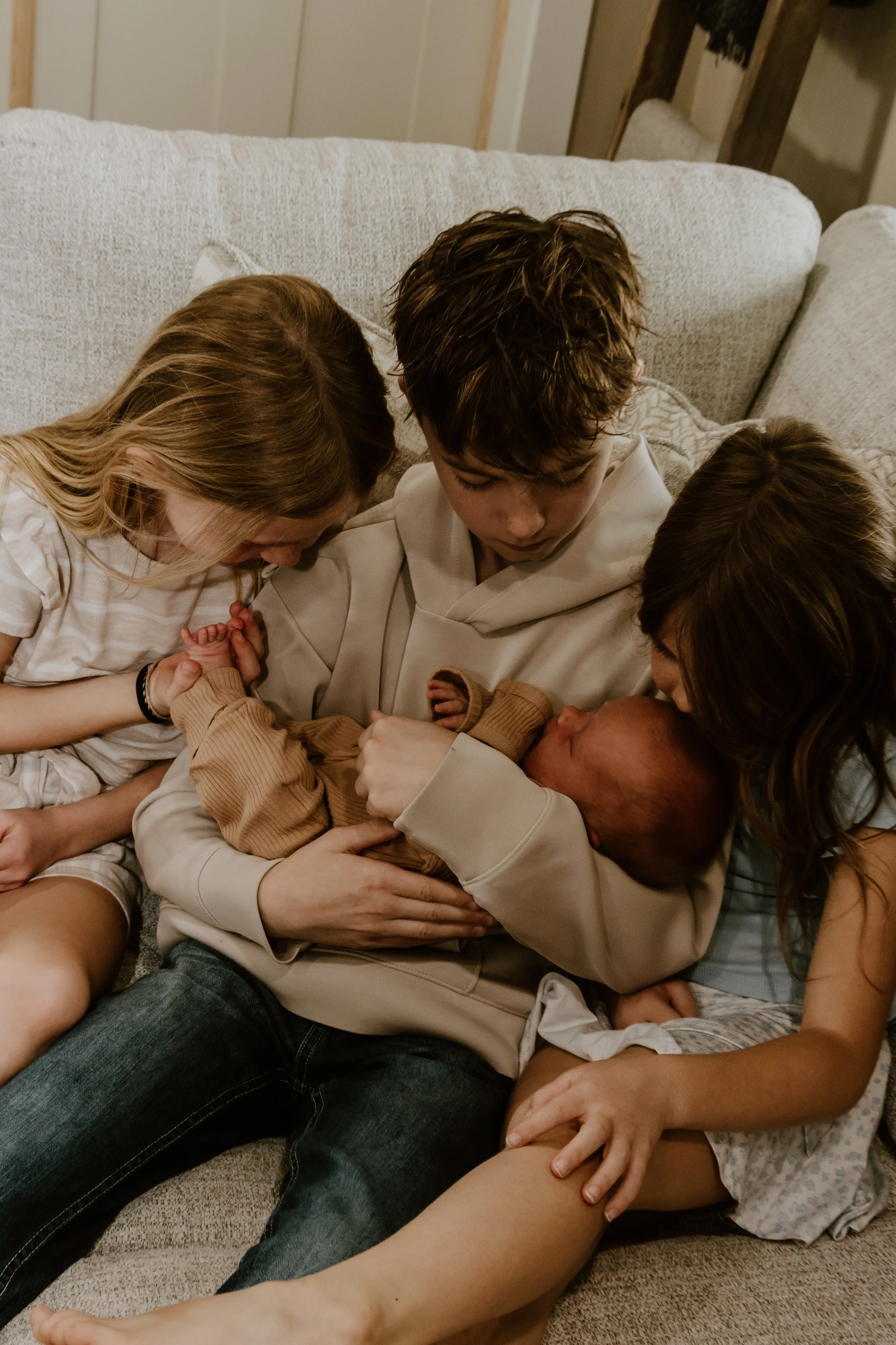 Children and adults gathered closely, holding a newborn baby, with everyone appearing to be focused on the infant in a cozy indoor setting.