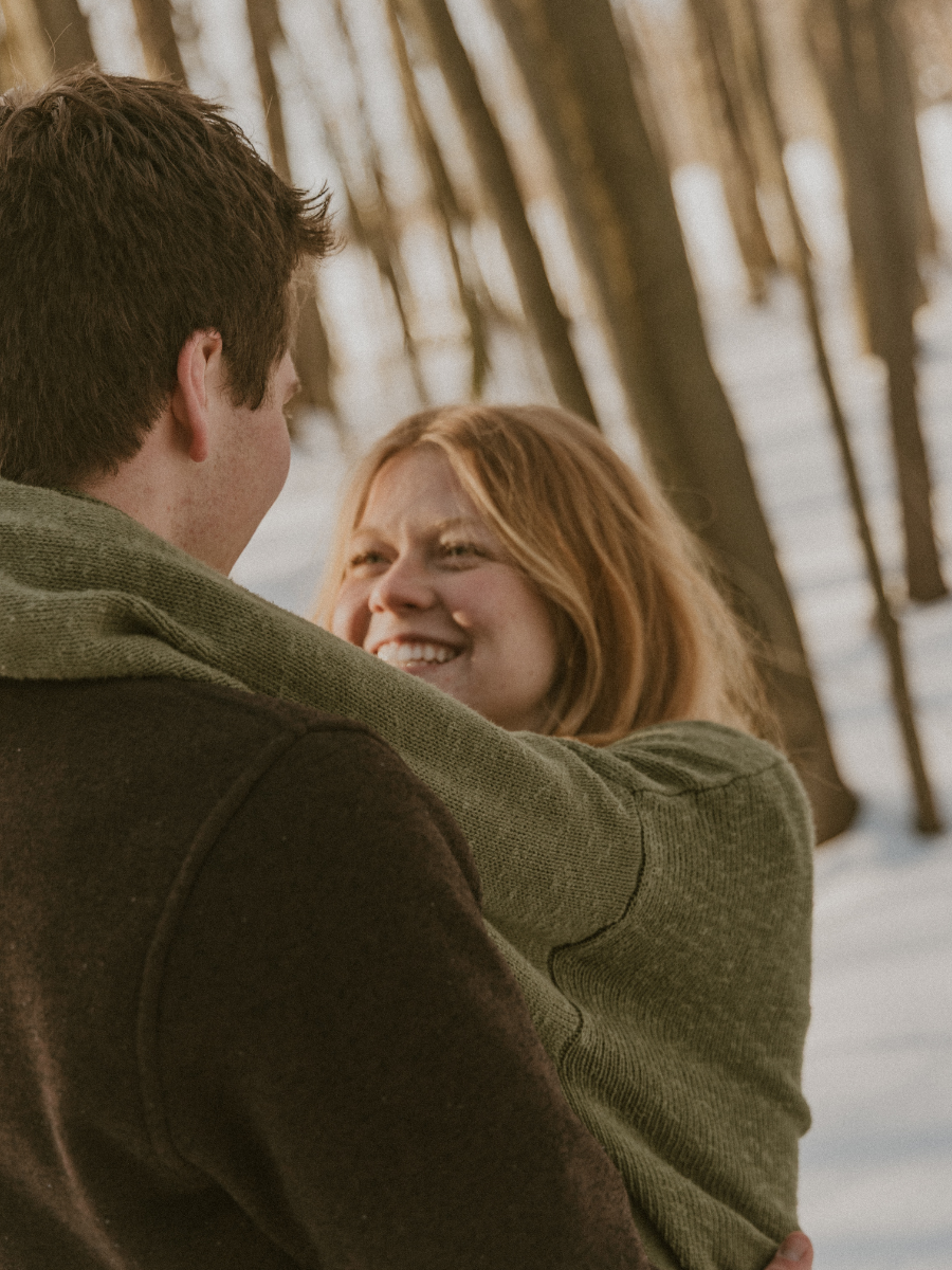 A young man and woman are embracing outdoors near a body of water with trees in the background. The woman is smiling and looking at the man.