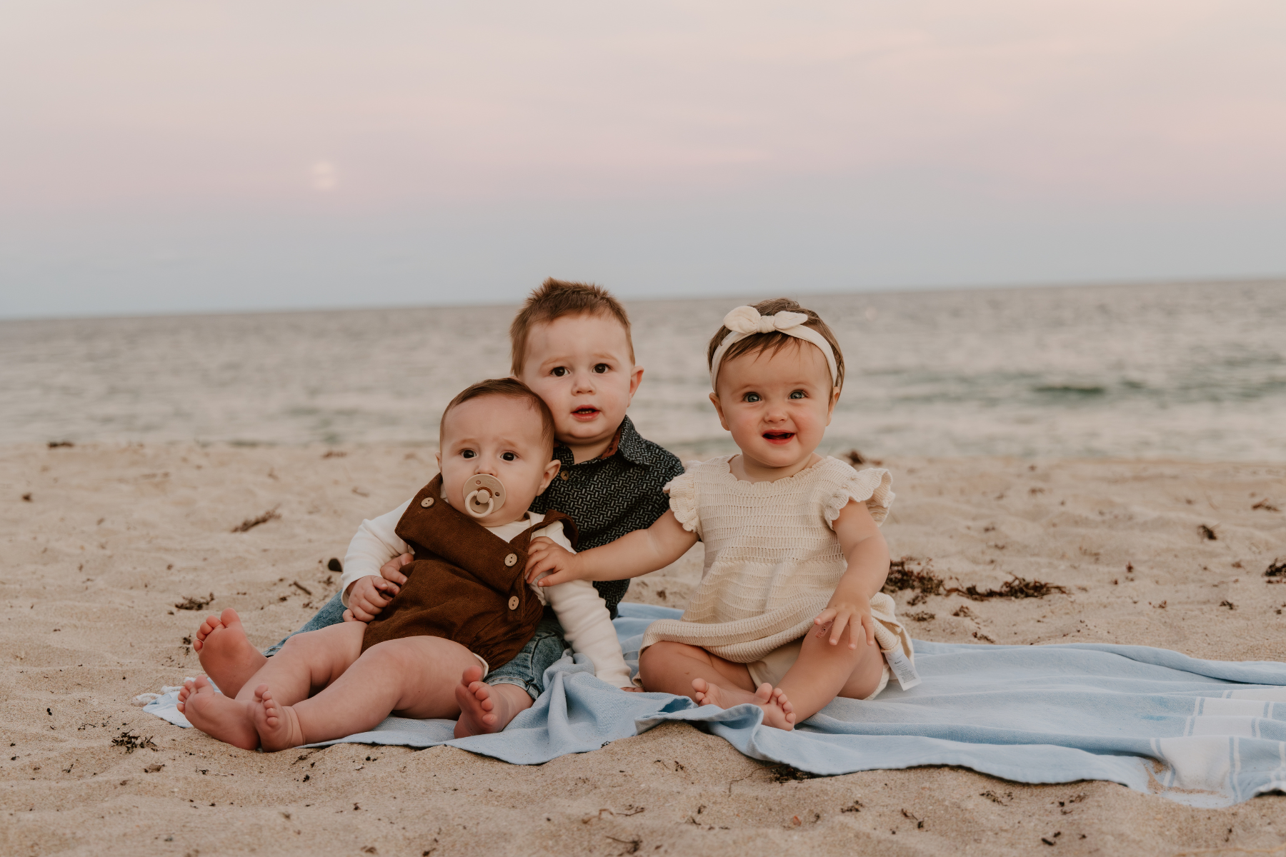 Three young children sitting on a blanket on a sandy beach, with the ocean and a pinkish sky in the background.