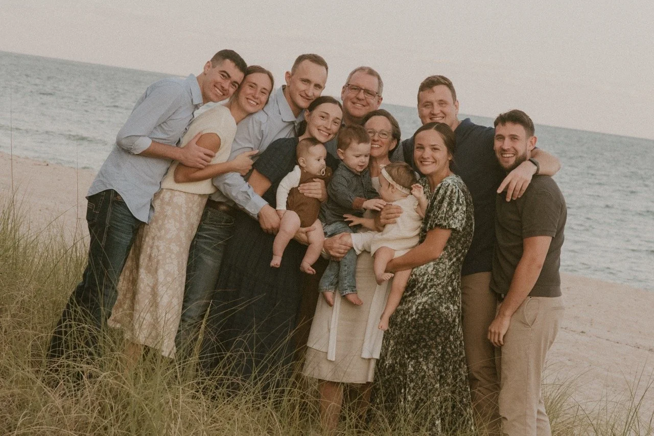 A large group of family and friends gathered on a beach, smiling and posing for a photo, with the ocean and sky in the background.