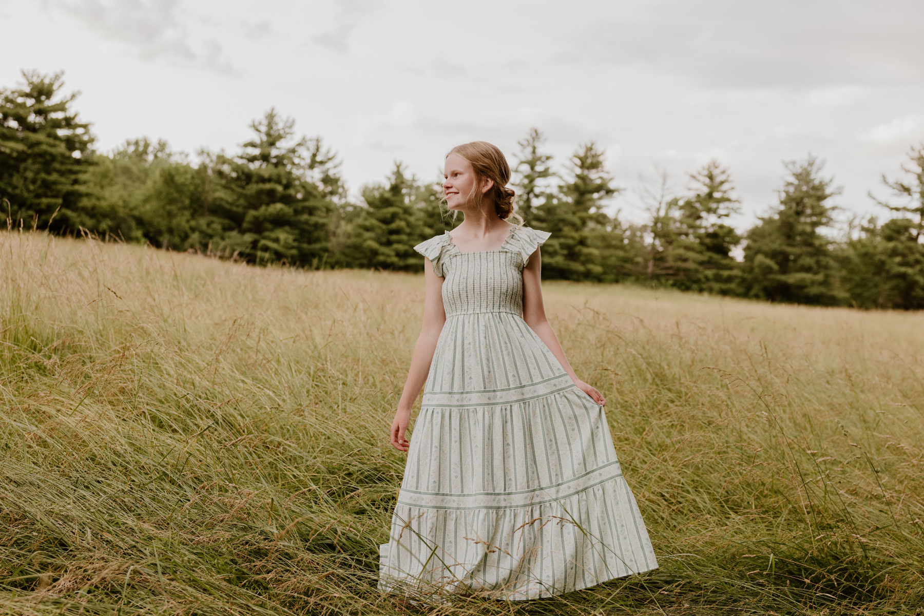 A young woman in a long, light-colored, striped dress standing in a grassy field with trees in the background and cloudy sky overhead.