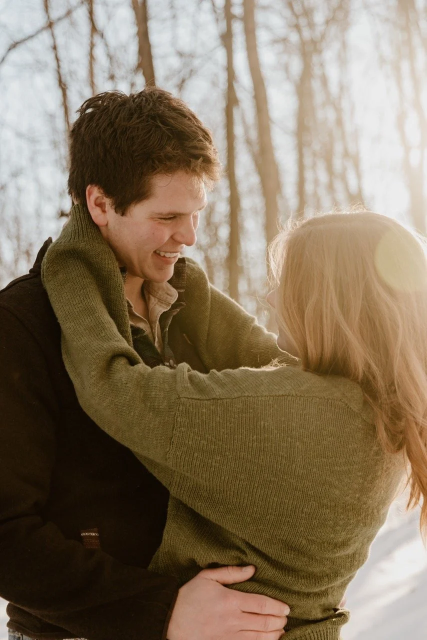 A young couple is outdoor in a wooded area, embracing and smiling at each other during daylight.
