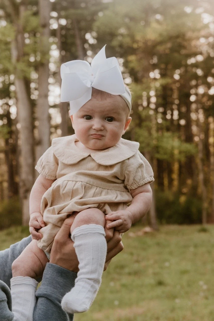 A baby girl with a large white bow on her head, wearing a beige dress and white socks, being held up outdoors in front of trees.