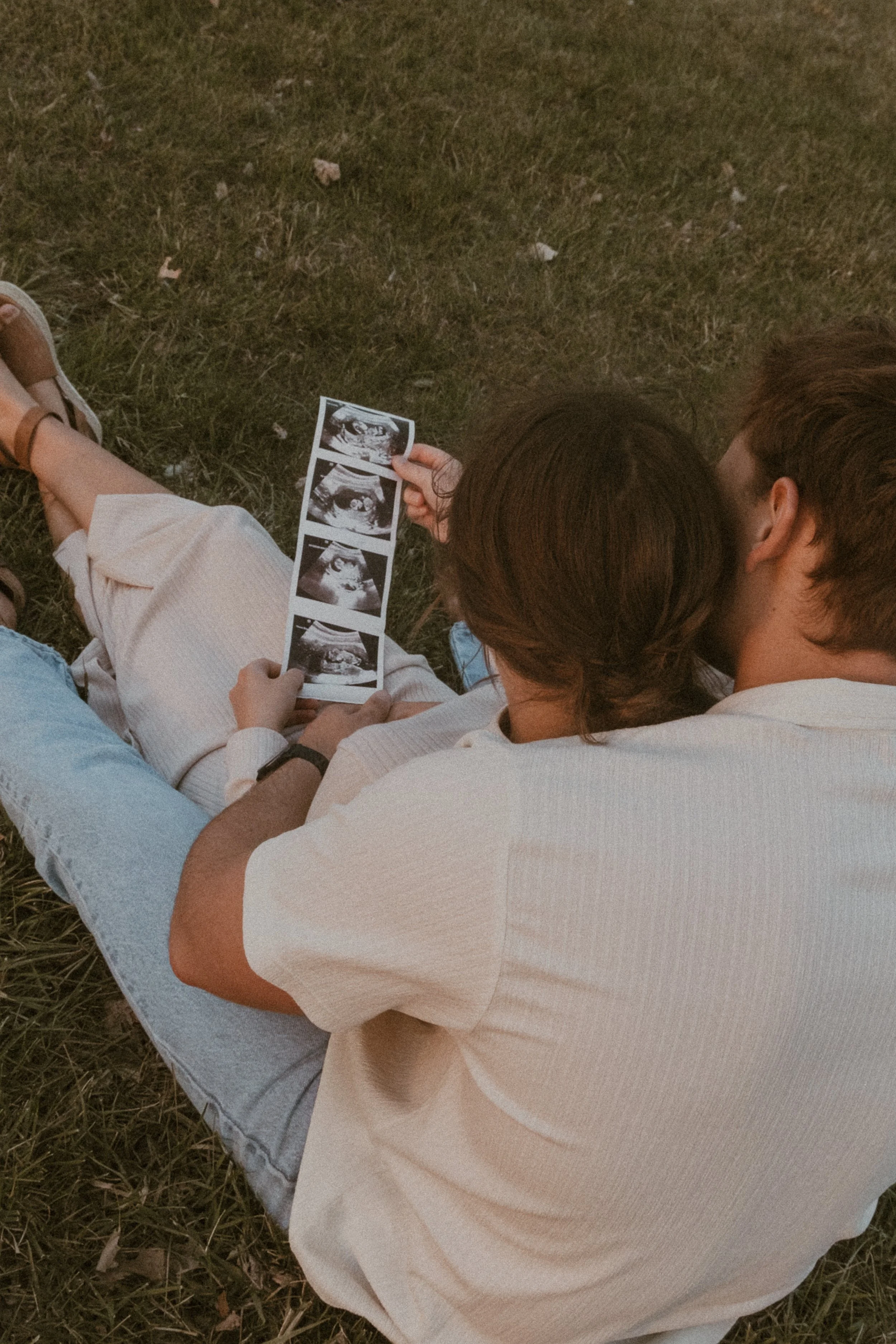 People sitting on the grass, looking at photo strips of images, with one person having brown hair and wearing a white shirt, and another with brown hair in a bun, wearing a white coat.