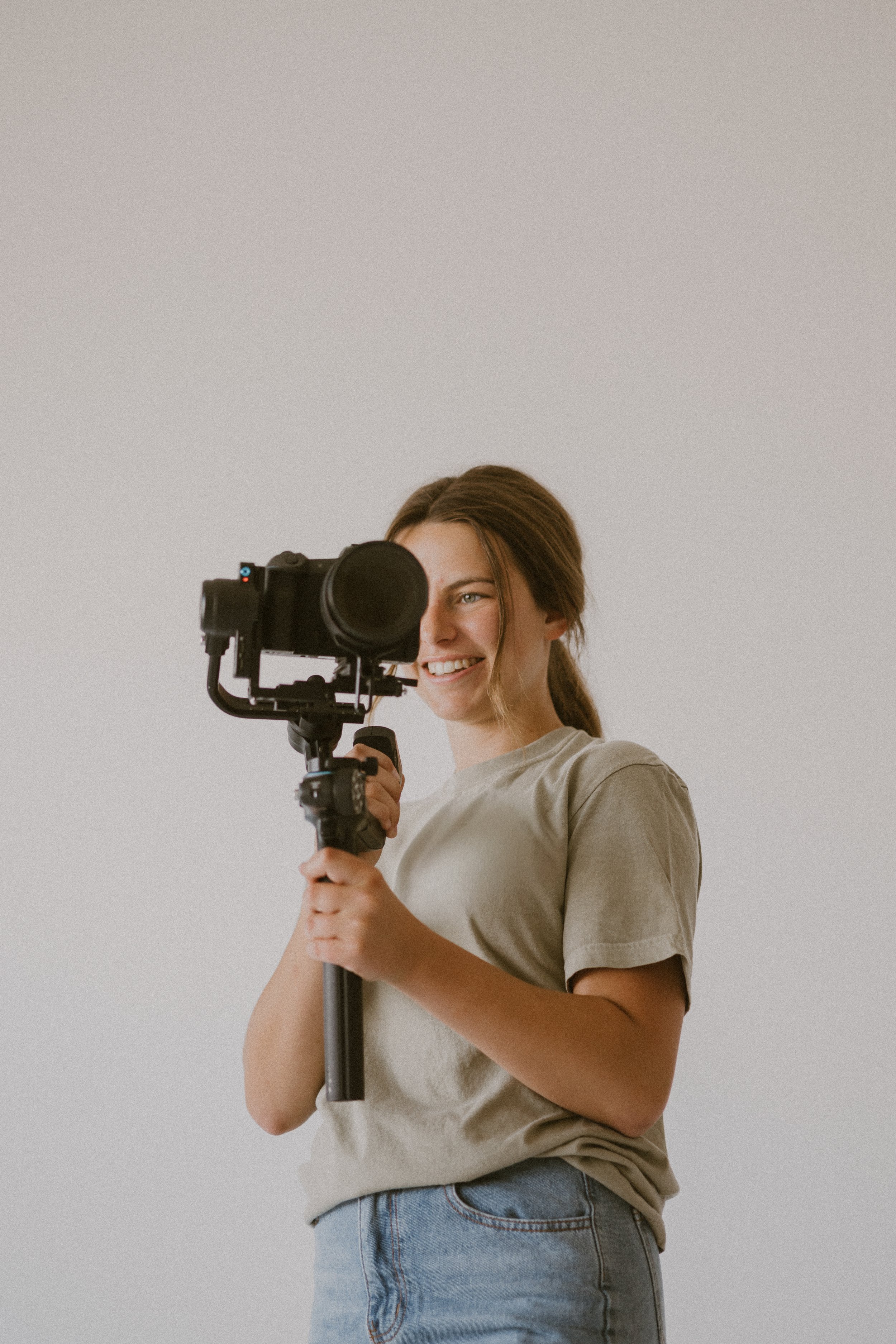 Young woman holding a camera on a tripod, smiling and looking at the camera, against a plain light-colored background.