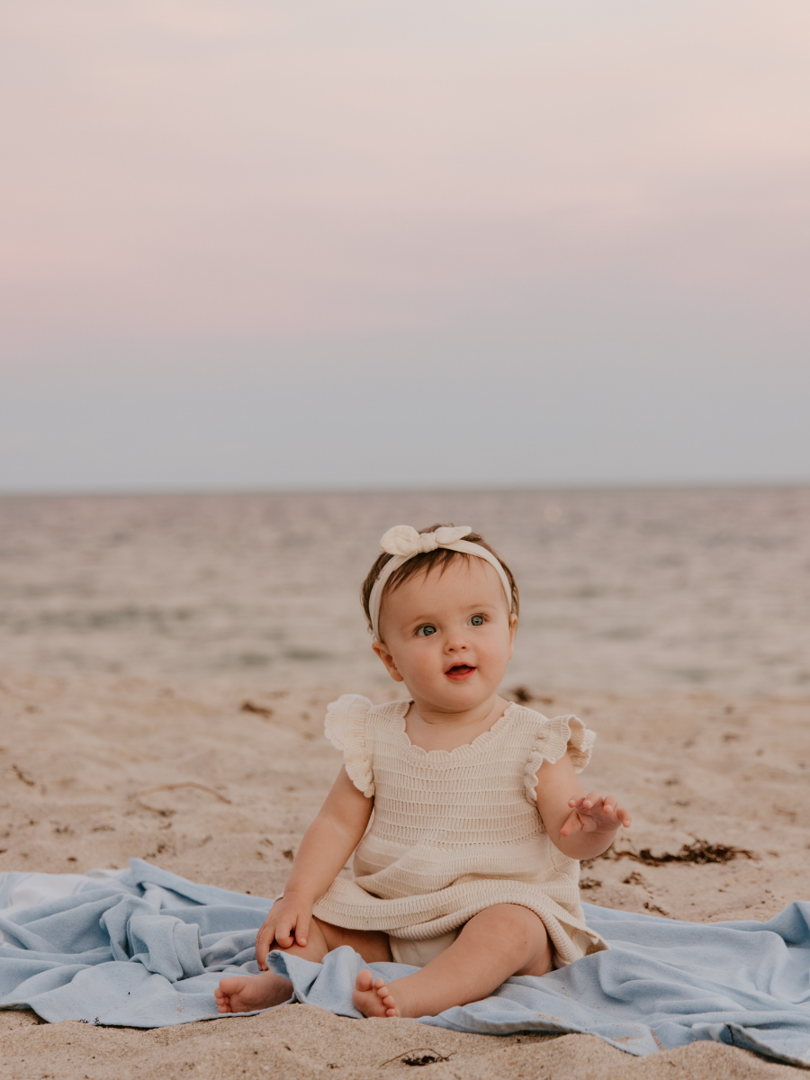 A young girl sitting on a blue blanket on the beach during sunset, wearing a cream-colored dress and a beige headband with a bow.