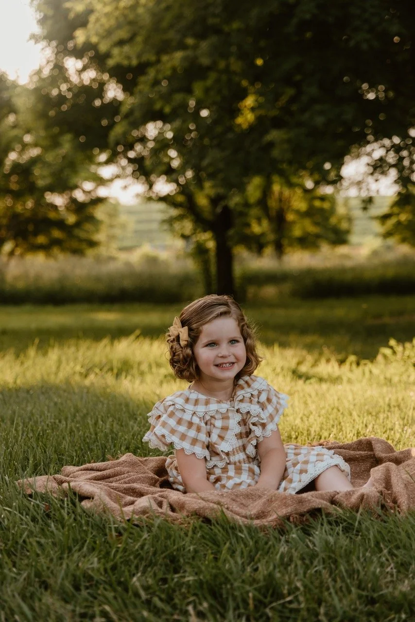 A young girl with curly hair and a tan bow sits on a blanket in a grassy park, smiling at the camera. Trees and sunlight are visible in the background.