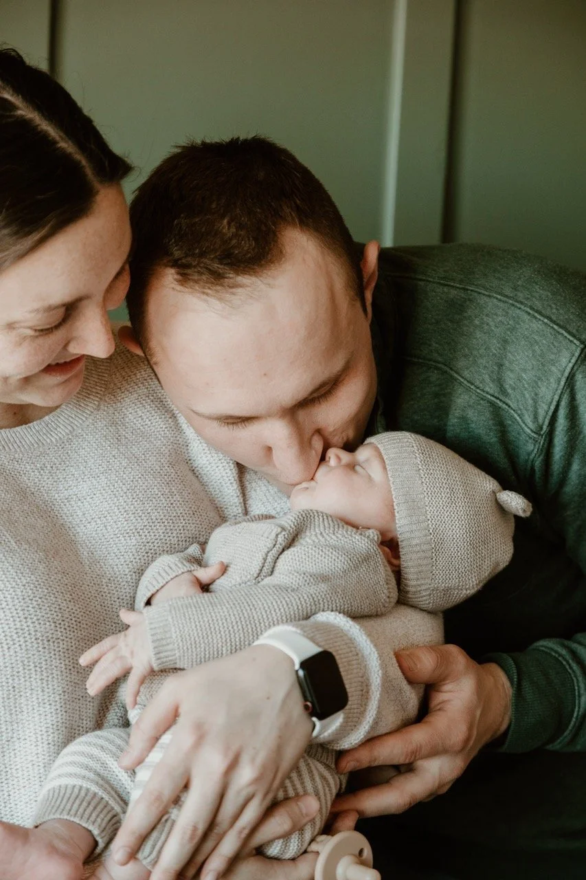 A family is holding their newborn baby. The father is kissing the baby's forehead, the mother is smiling and holding the baby, and the baby is wearing a beige hat and sweater.