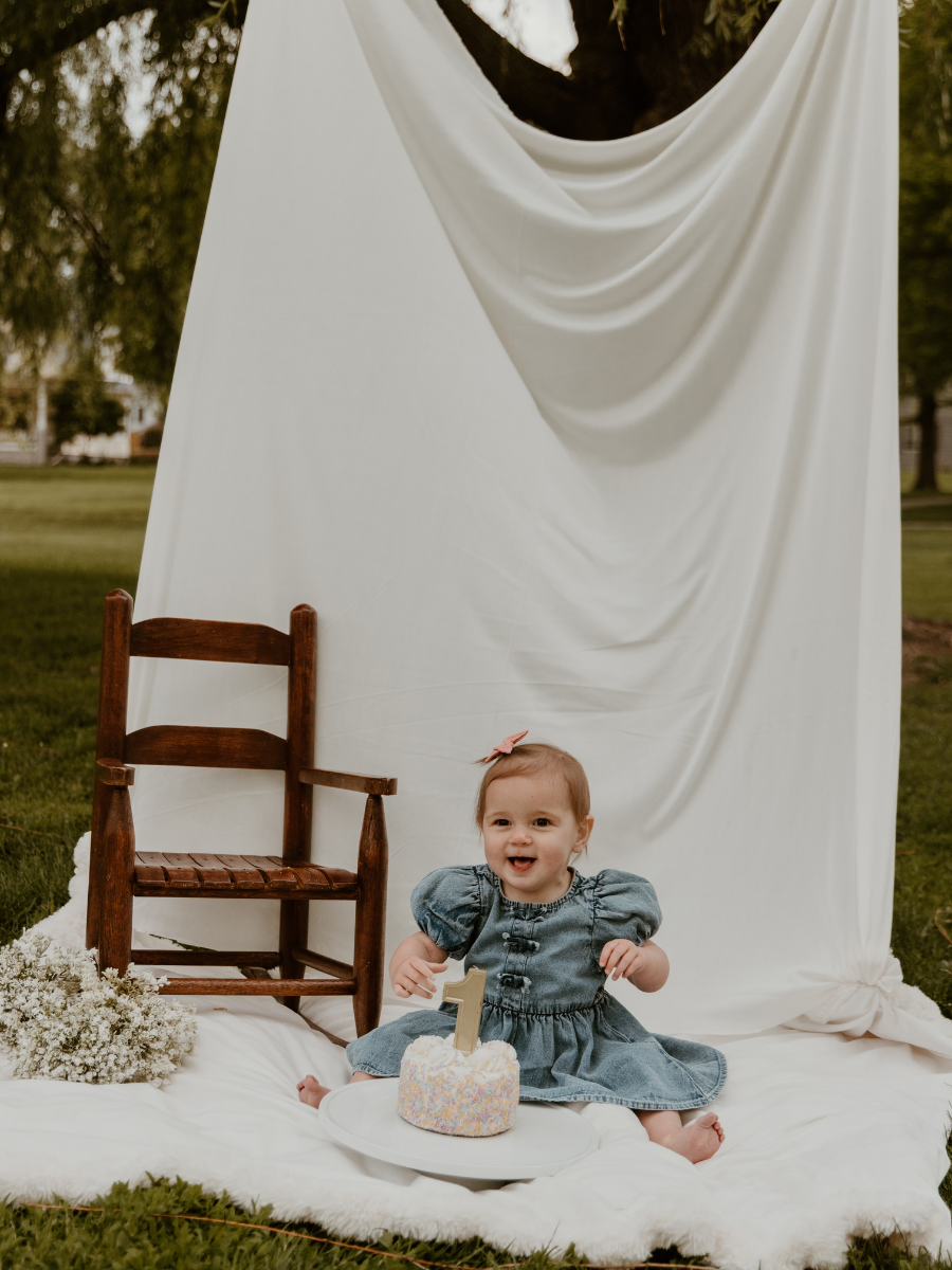 A baby girl sitting on a white blanket outside, celebrating her first birthday with a small cake that has a number one candle. There is a wooden chair and a floral arrangement beside her, with a white draped backdrop hanging from a tree in the background.