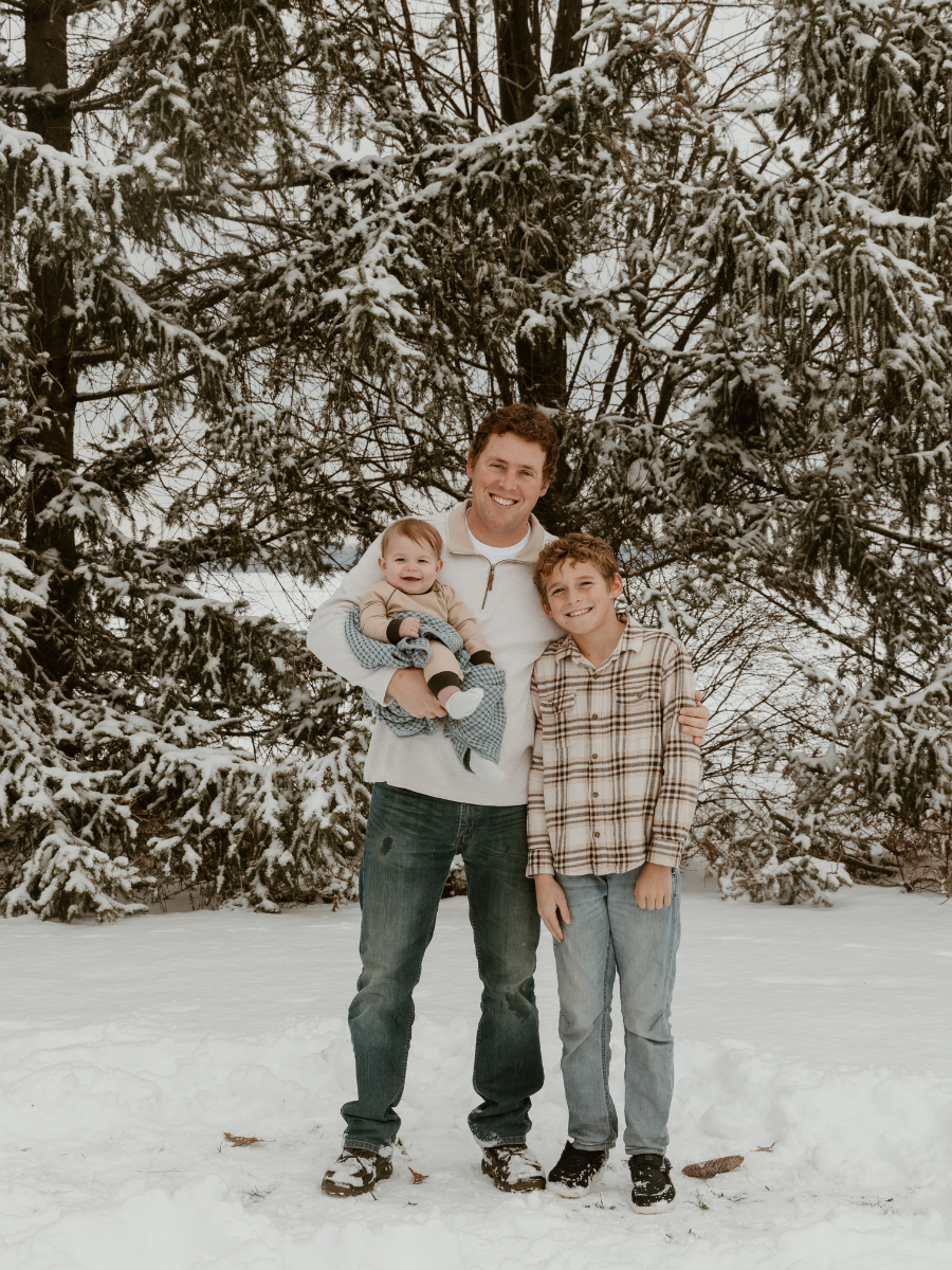 A man and two children standing in snow in front of snow-covered trees. The man is holding a baby, and the older child is standing next to them.