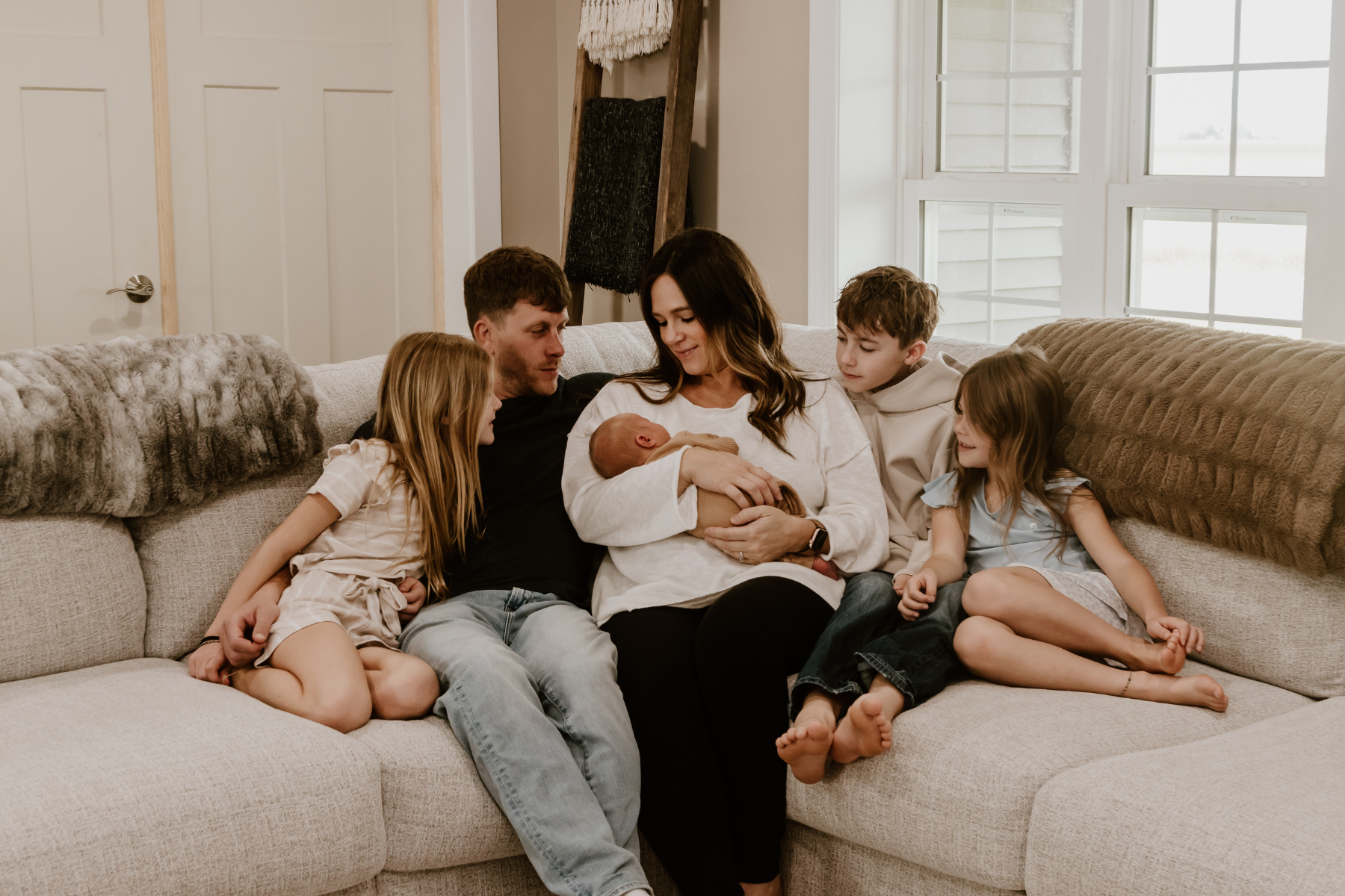 A family of six sitting together on a light-colored sectional sofa in a living room. The woman is holding a newborn baby, and the rest of the children surround them, all looking at the baby.