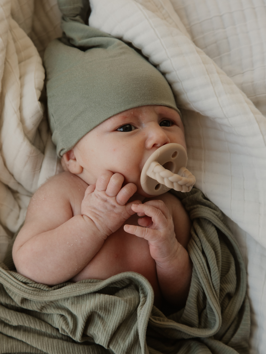 A baby with a pacifier in their mouth, lying in bed with a gray hat and striped clothing, resting on white blankets.