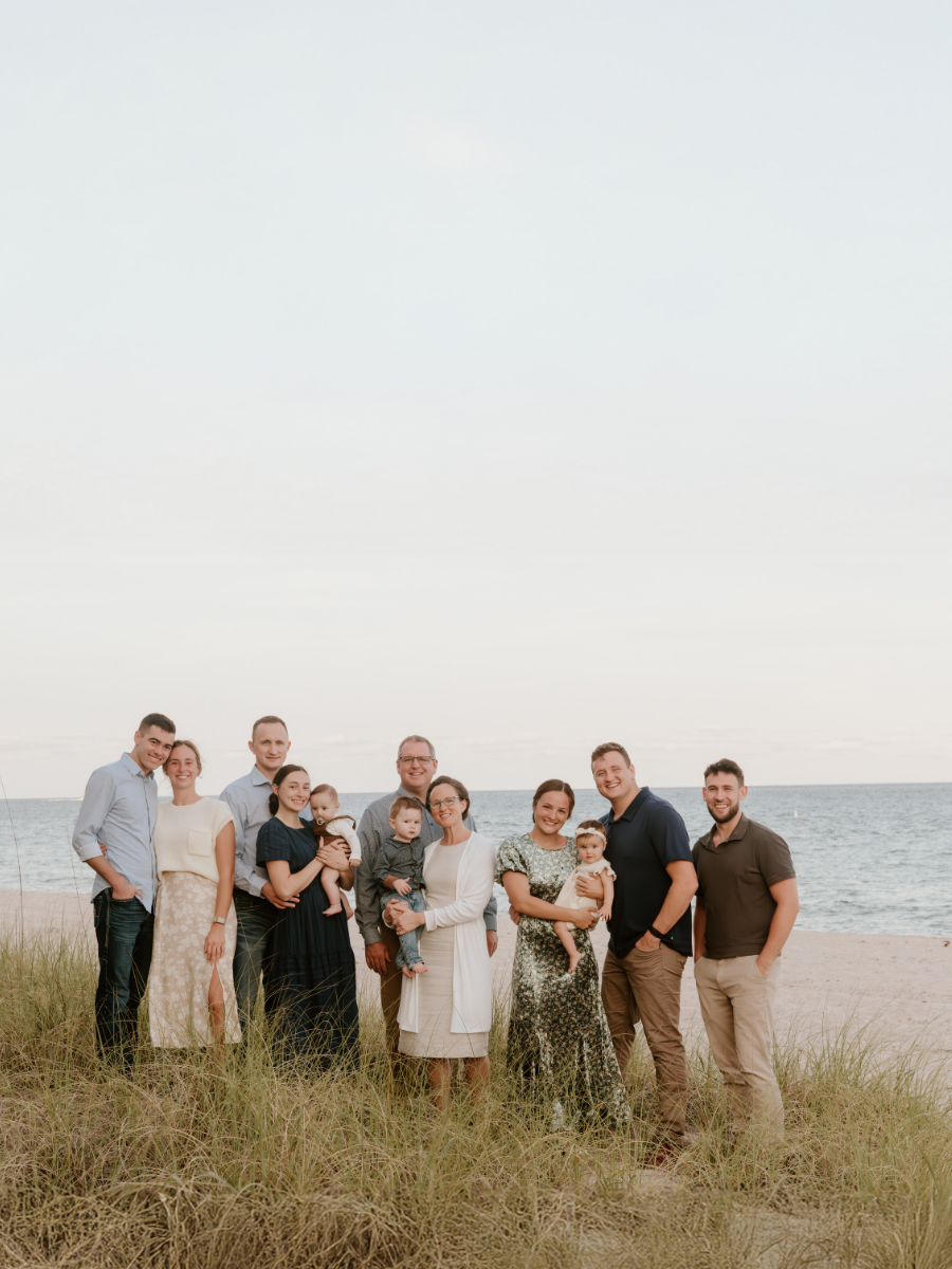 Family photo on the beach with adults and children standing on sand and grass, ocean in the background, during daytime, smiling and enjoying the visit.