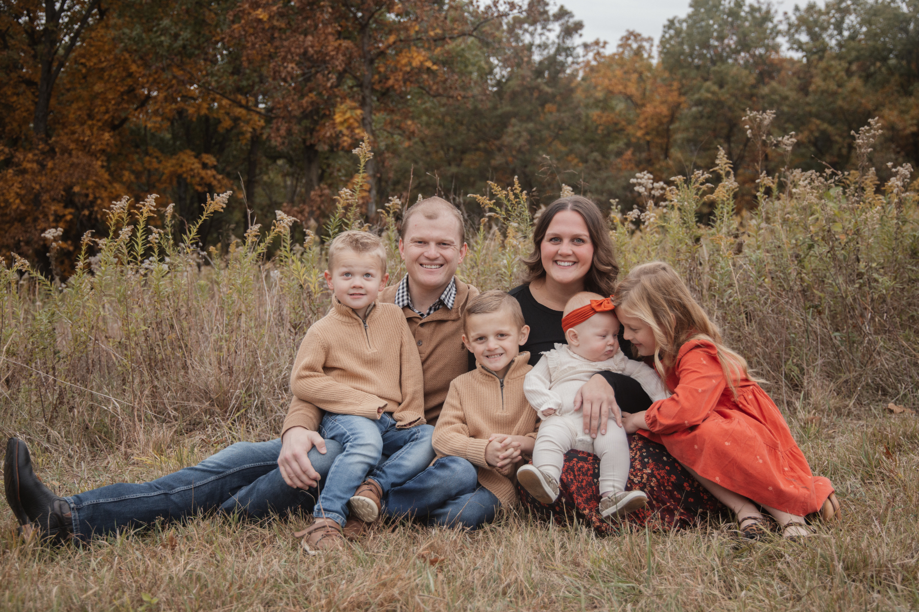 A family of six sitting in a grassy field with fall foliage in the background. The family includes a father, mother, and four children. The children are wearing autumn-colored clothes, and the family is smiling and enjoying the outdoor setting.