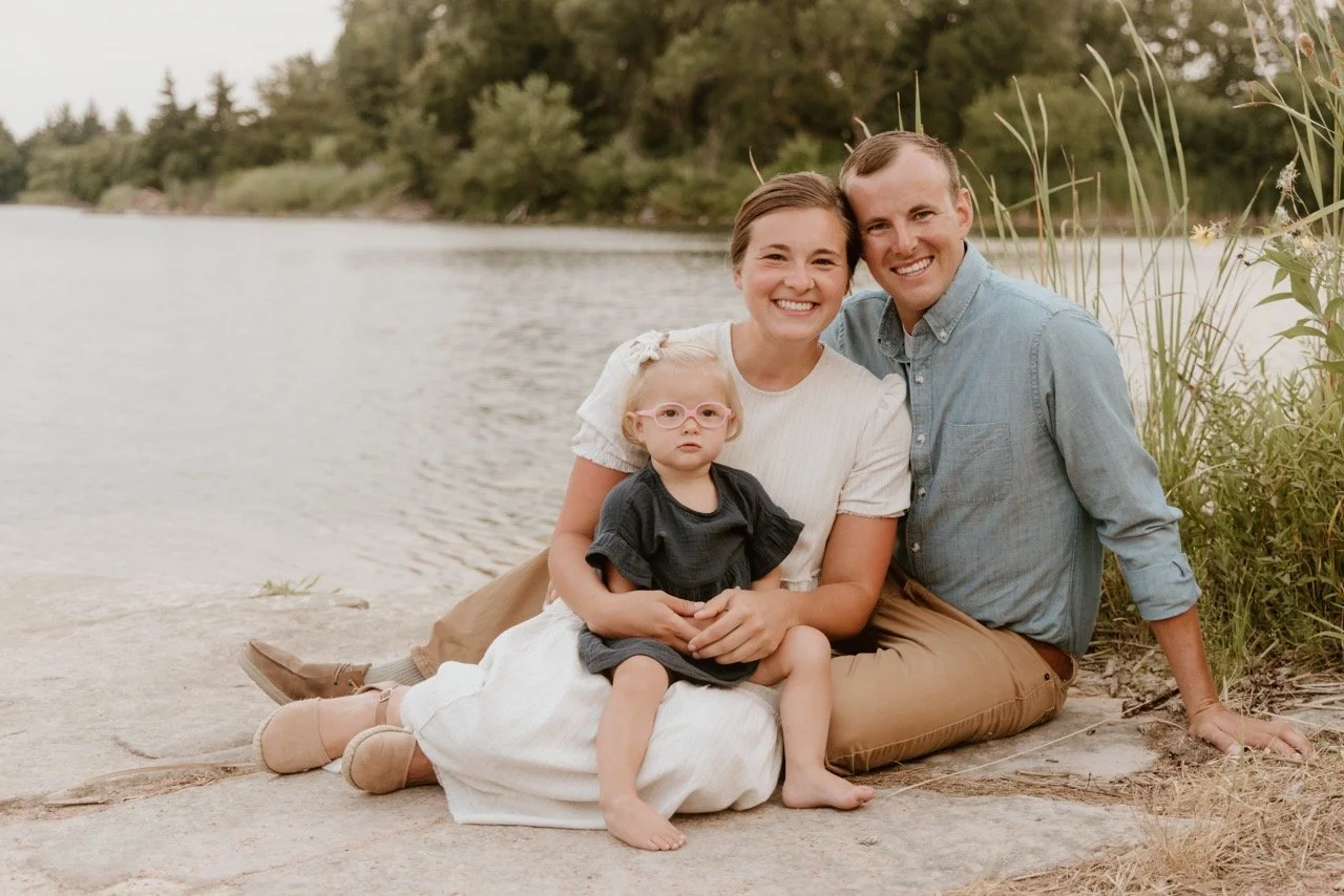 A happy family of three sitting on a riverbank, smiling and enjoying outdoor time. The family includes a young girl with glasses, her mother, and her father. The background features water, trees, and greenery.