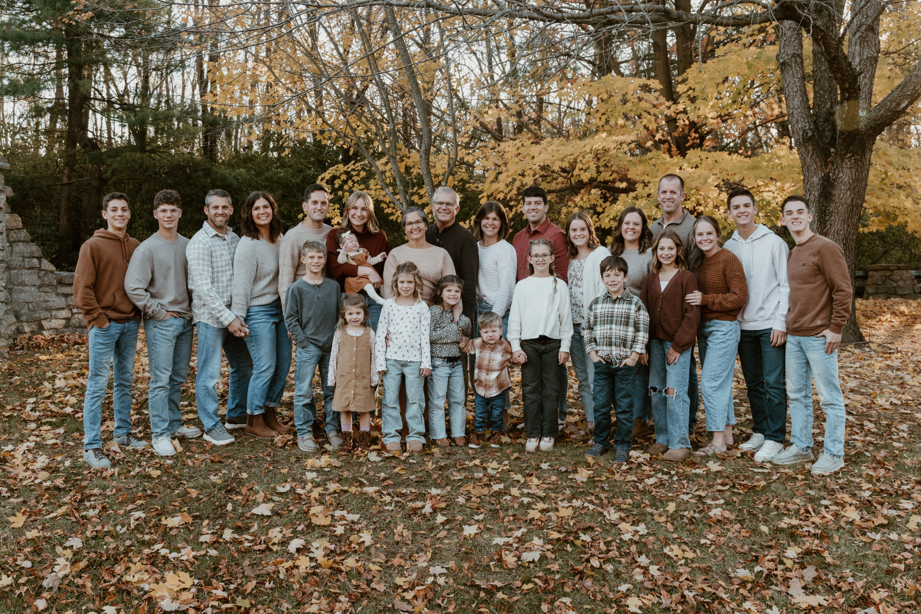 Family photo of adults and children outdoors during autumn, standing on fallen leaves with trees and a stone wall in the background.