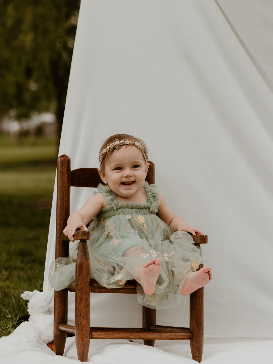 A smiling baby girl in a green dress with floral embroidery, sitting on a wooden chair outdoors in front of a white backdrop.