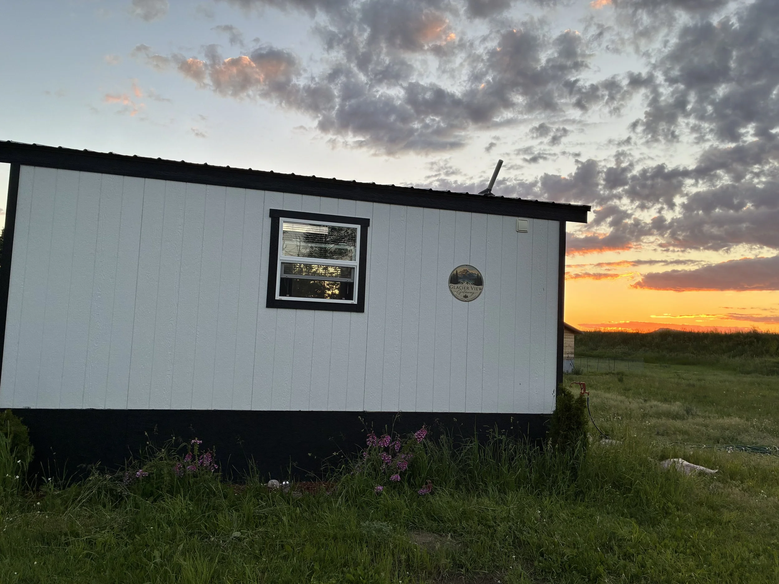 A building with white vertical siding and black trim around the window and roof at sunset, with a landscape of green grass and purple flowers in the foreground and a sky filled with dark and pink clouds.