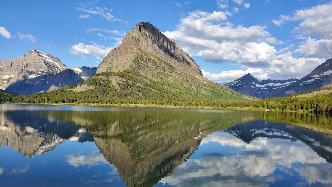 Scenic landscape of a mountain reflected in a calm lake, with a blue sky and scattered clouds.