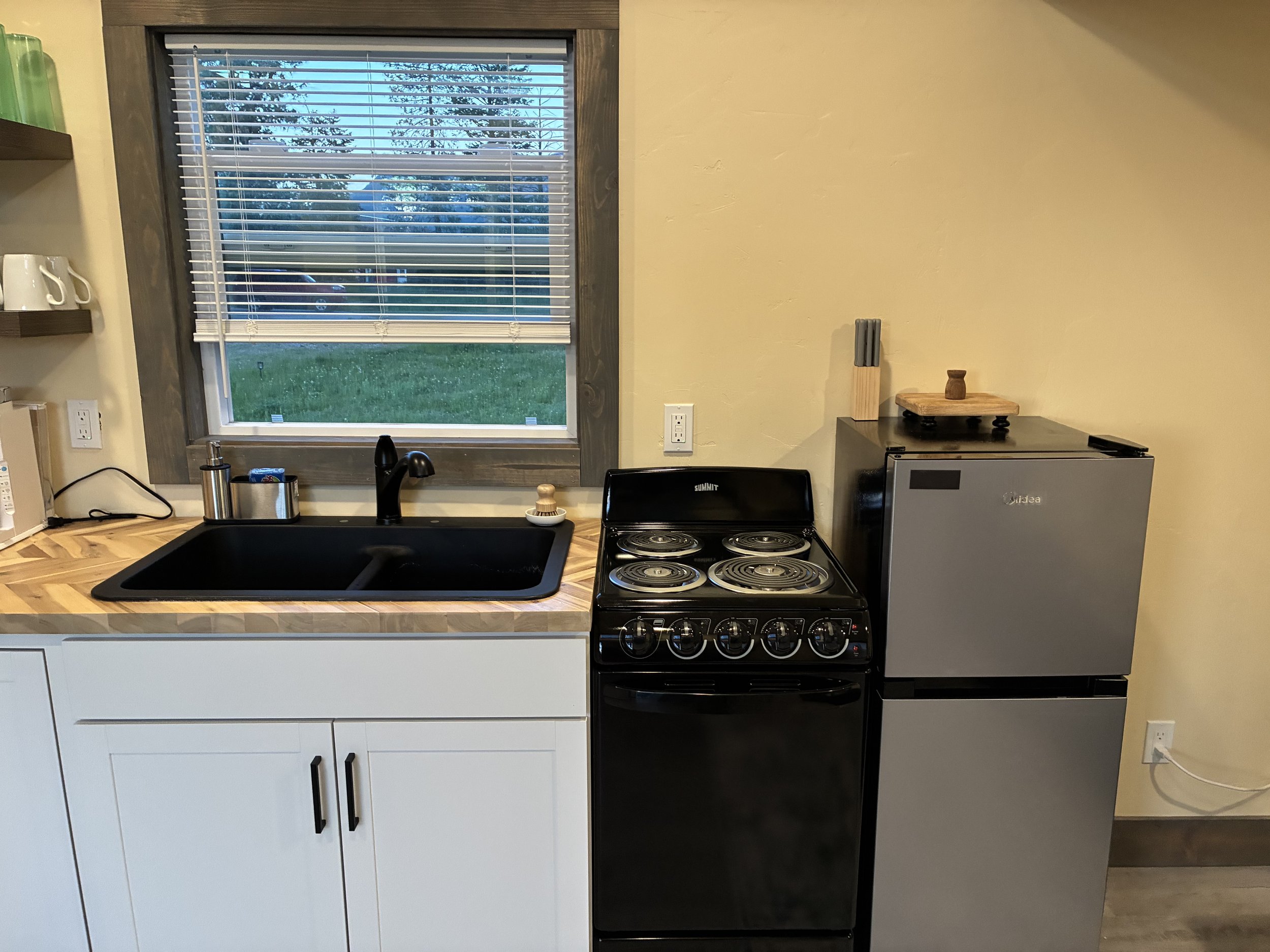 Kitchen with a window showing trees outside, a black sink, a small stovetop, a mini fridge, a cutting board with a small wooden object and a bundle of knives, and two white shelves with cups and green glasses.