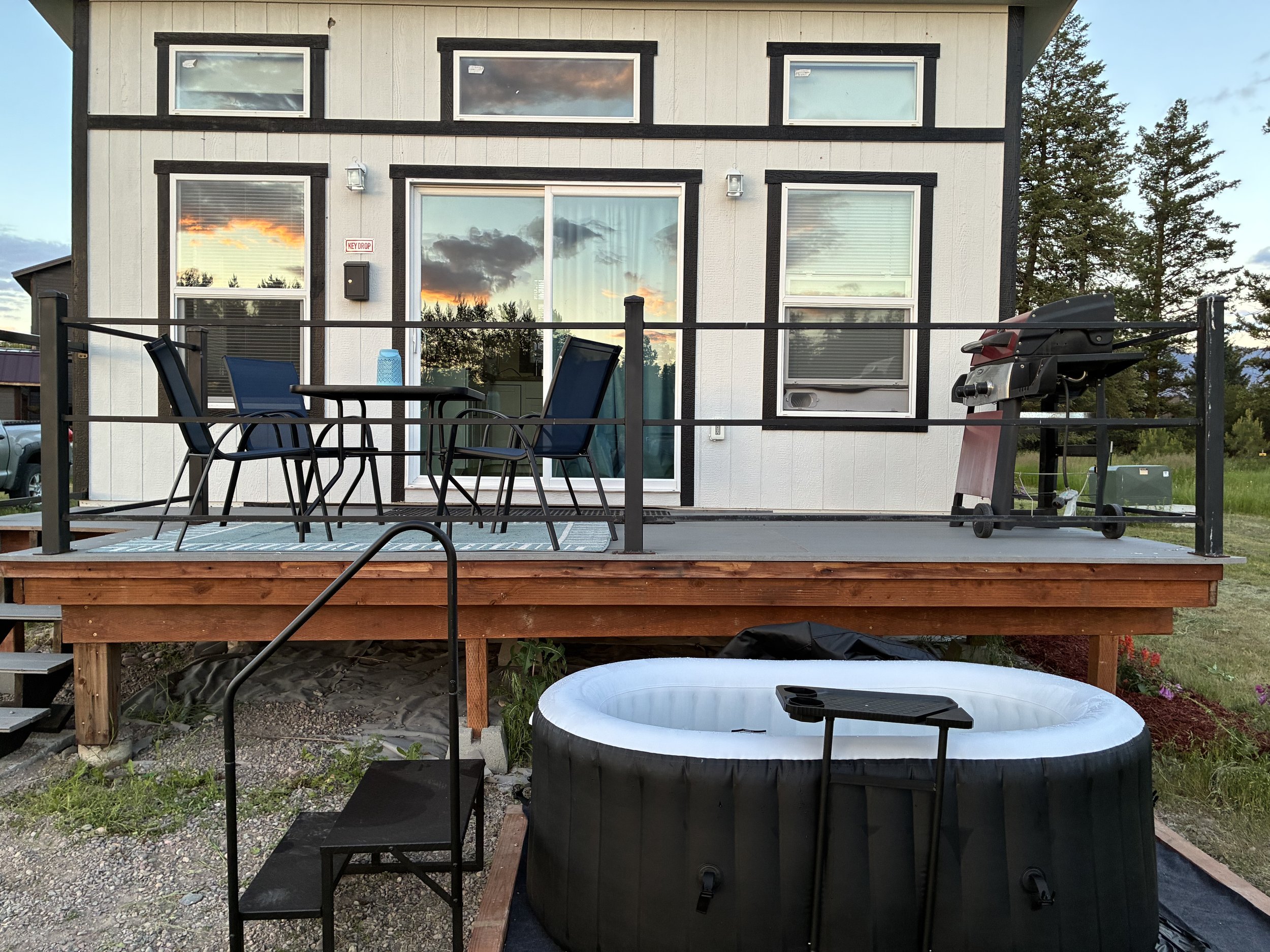 View of the back of a house with a wooden deck, outdoor furniture, a barbecue grill, a small inflatable pool, and a hot tub. The house has large windows reflecting a colorful sunset sky.