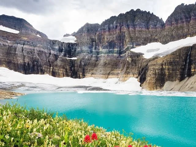 A vibrant turquoise lake surrounded by snow-capped mountains and green grass with colorful flowers in the foreground.