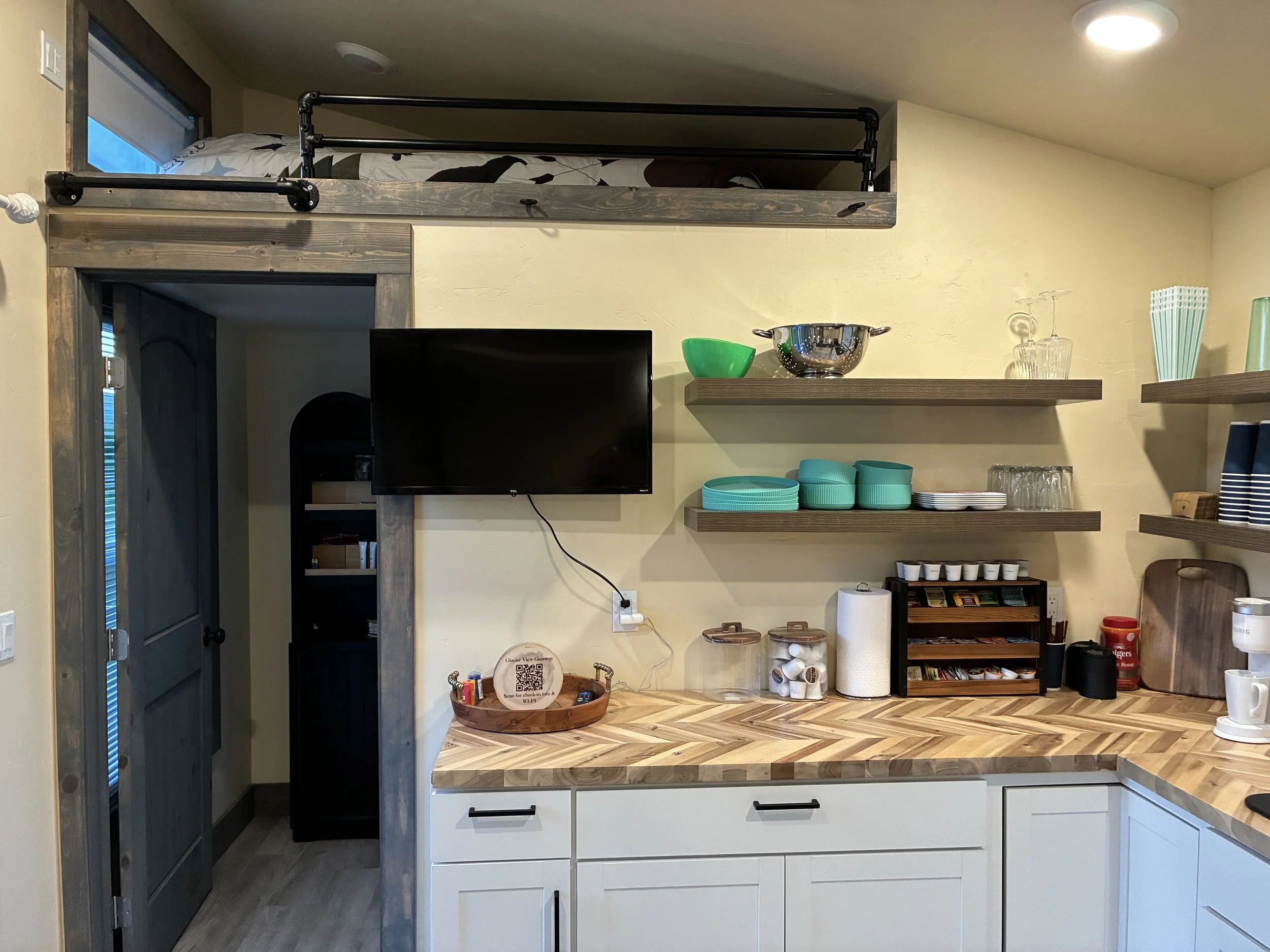A kitchen with a lofted bed above a countertop, a television, open shelves with dishes, and various kitchen items on the counter.