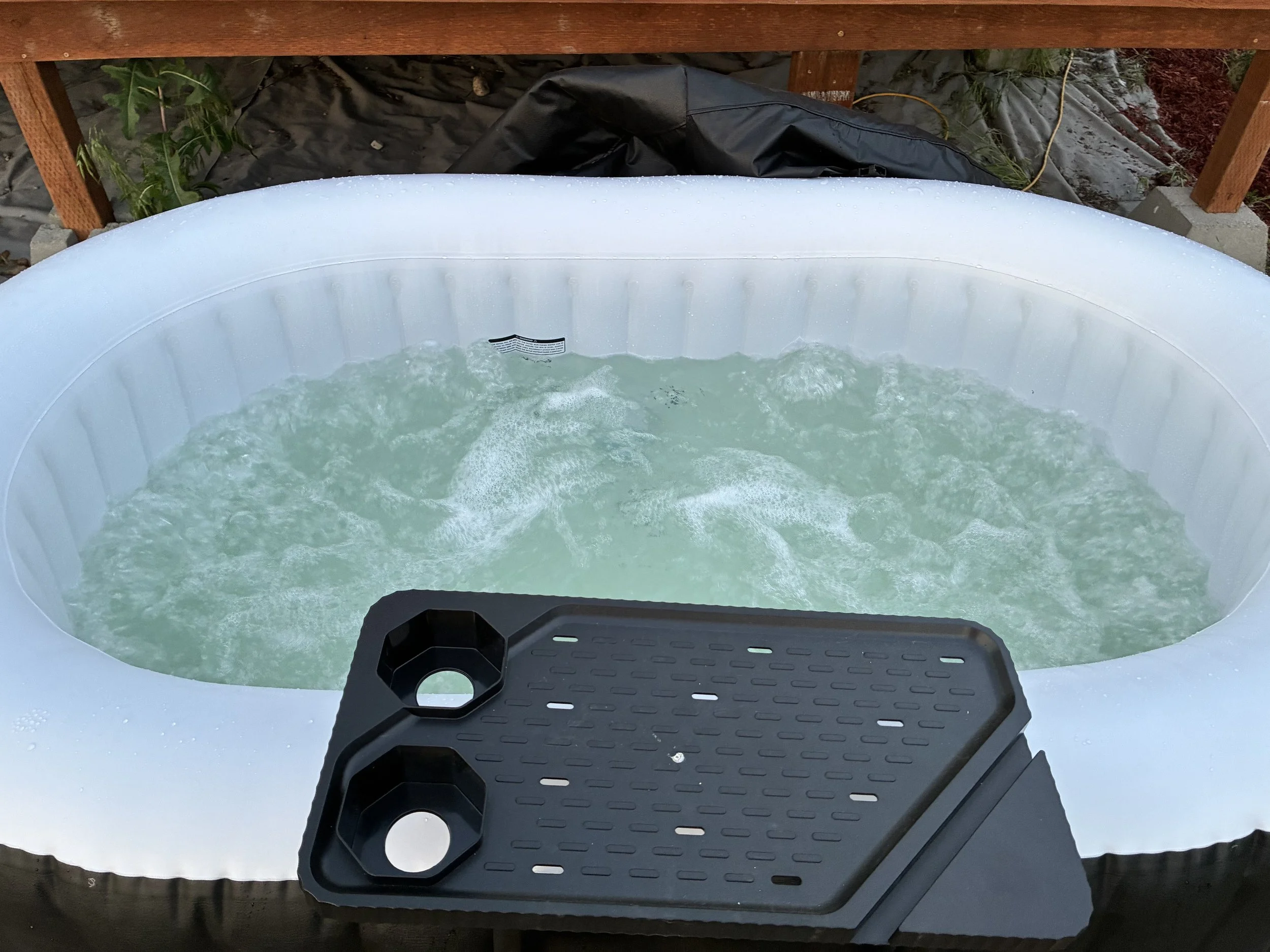 Inflatable hot tub filled with water, positioned outdoors on gravel, with a black tray on the edge, surrounded by a wooden deck, greenery, and garden surroundings.